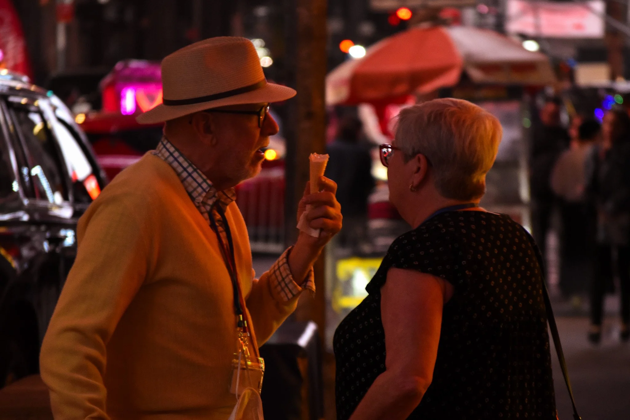 Two people talking outdoors at night, one holding an ice cream cone, surrounded by blurred city lights and umbrellas.