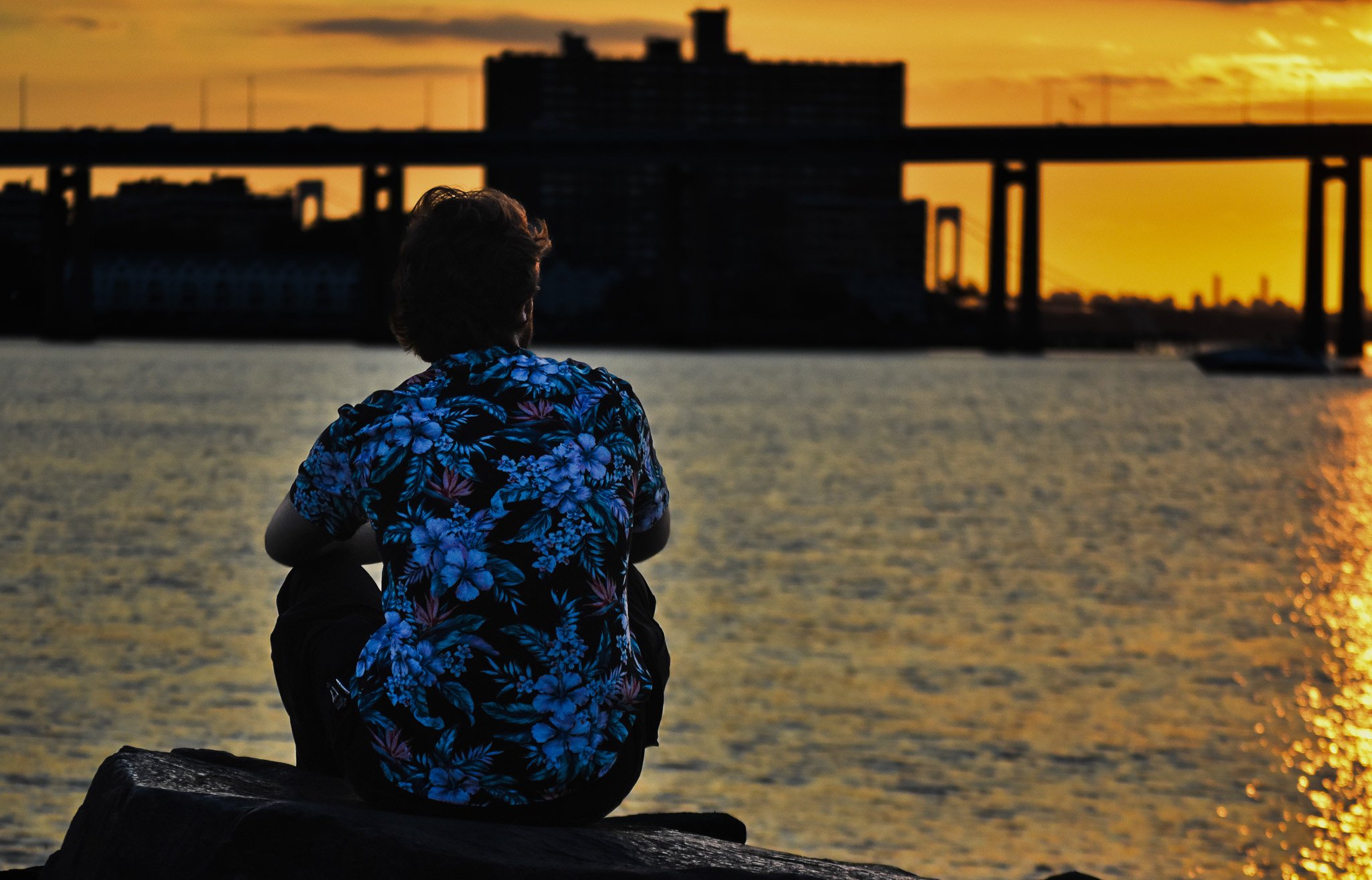 A person wearing a blue floral shirt sitting on a rock by the water, watching a sunset with a bridge and city skyline in the background.