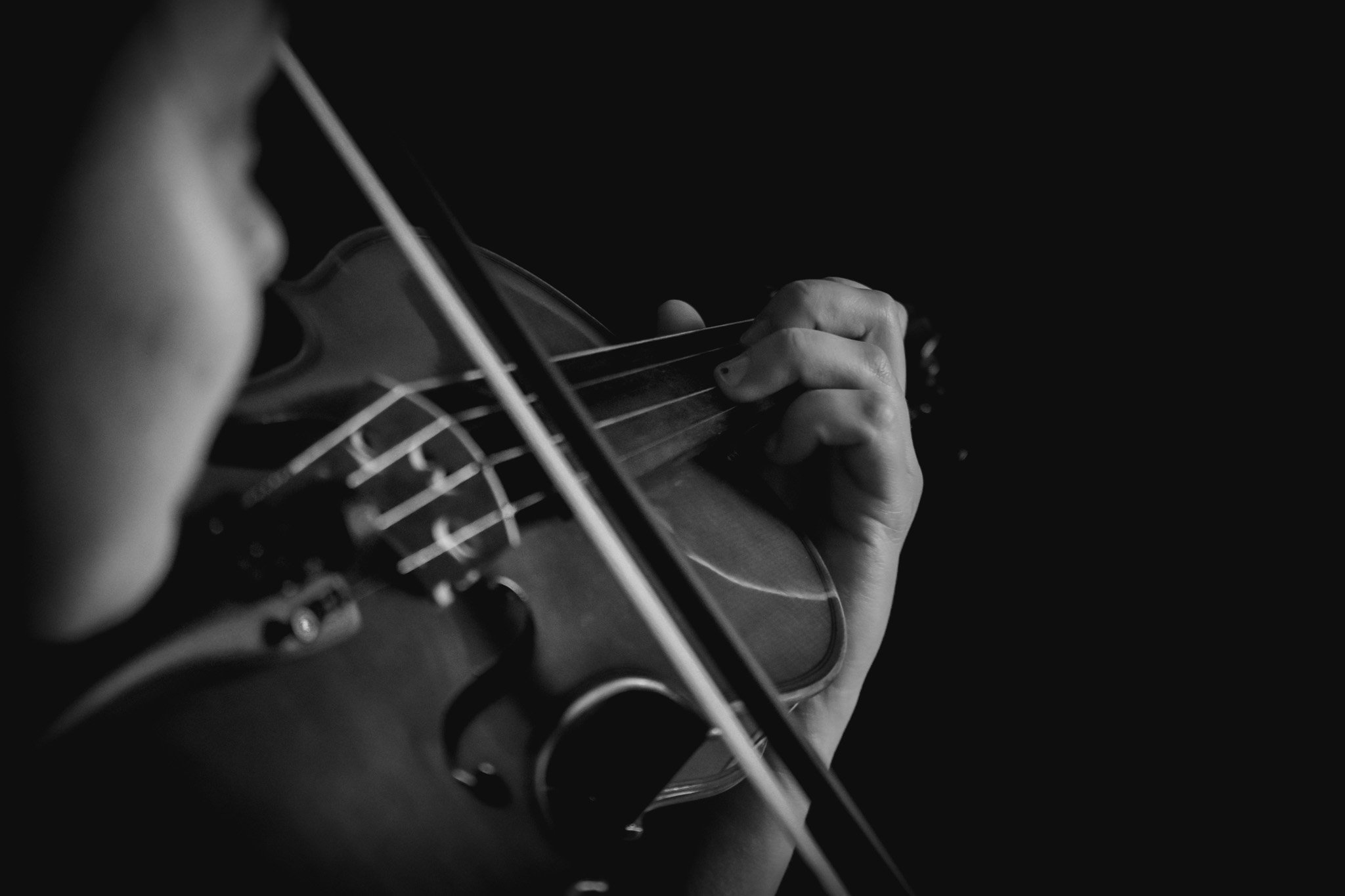 Close-up black and white photo of a person playing the violin, focusing on their hand pressing down the strings and part of their face blurred out.