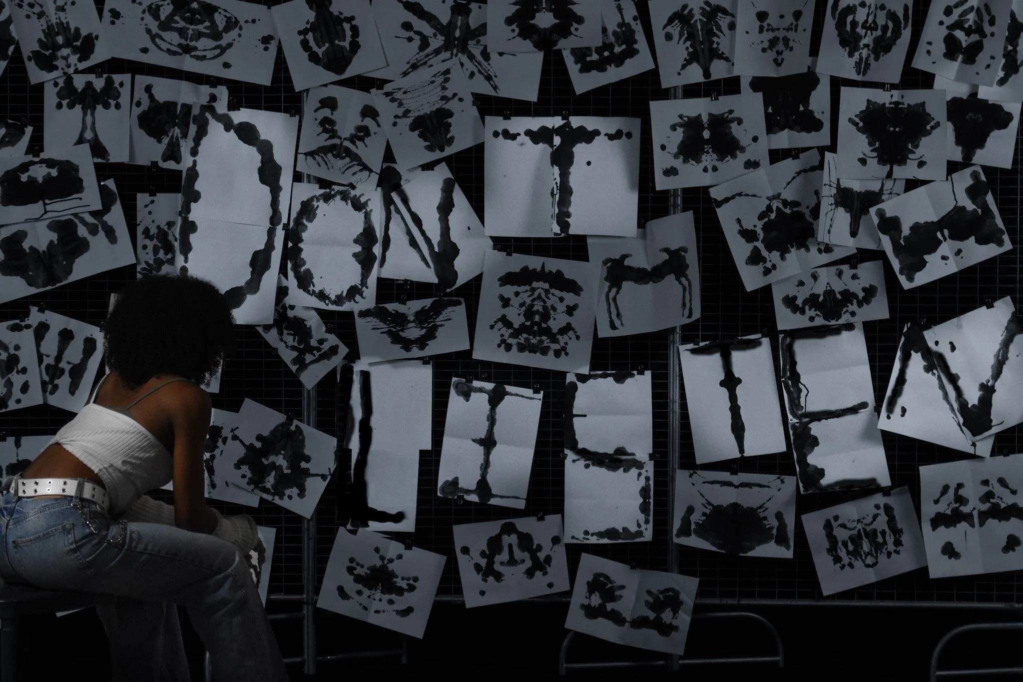 A person sitting and observing black inkblots on white papers pinned on a black grid wall, with various inkblot patterns displayed.