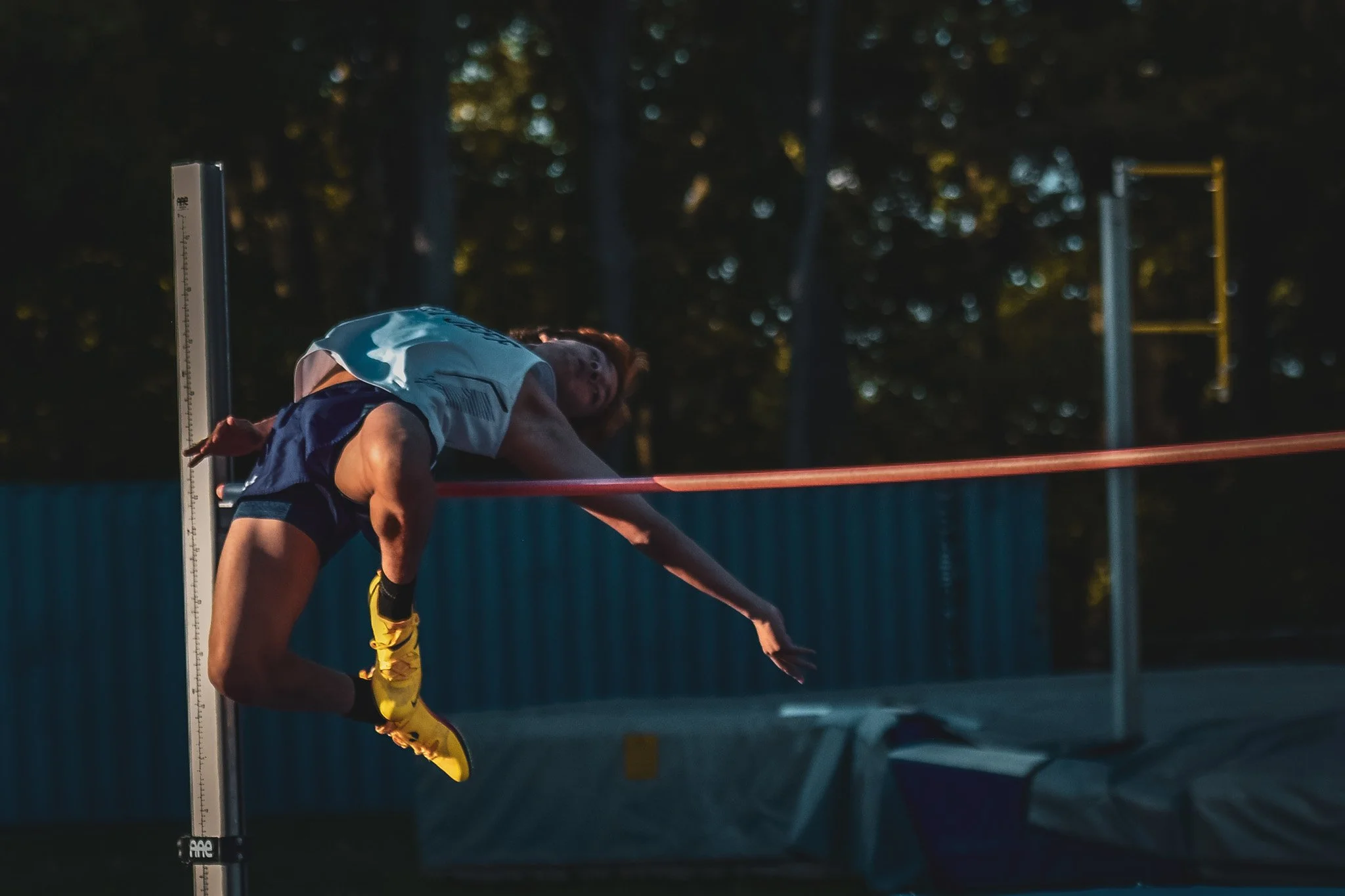 An athlete in a track and field event leaning forward with one hand touching the ground, near a measure at the starting point of a race, with trees in the background during evening or early morning.