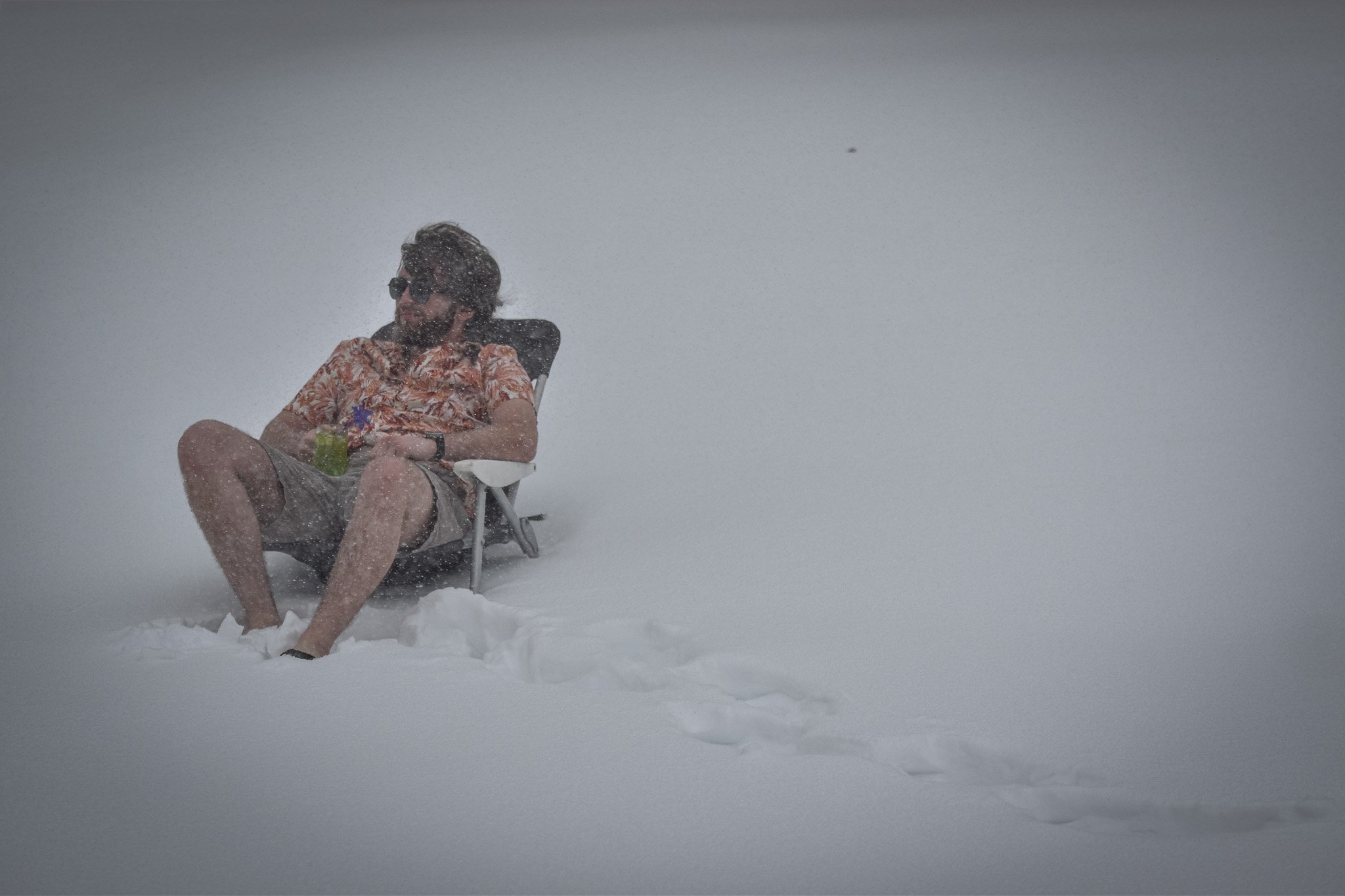 A man sitting in a chair in the snow, wearing sunglasses and a colorful Hawaiian shirt, with snow falling around him.
