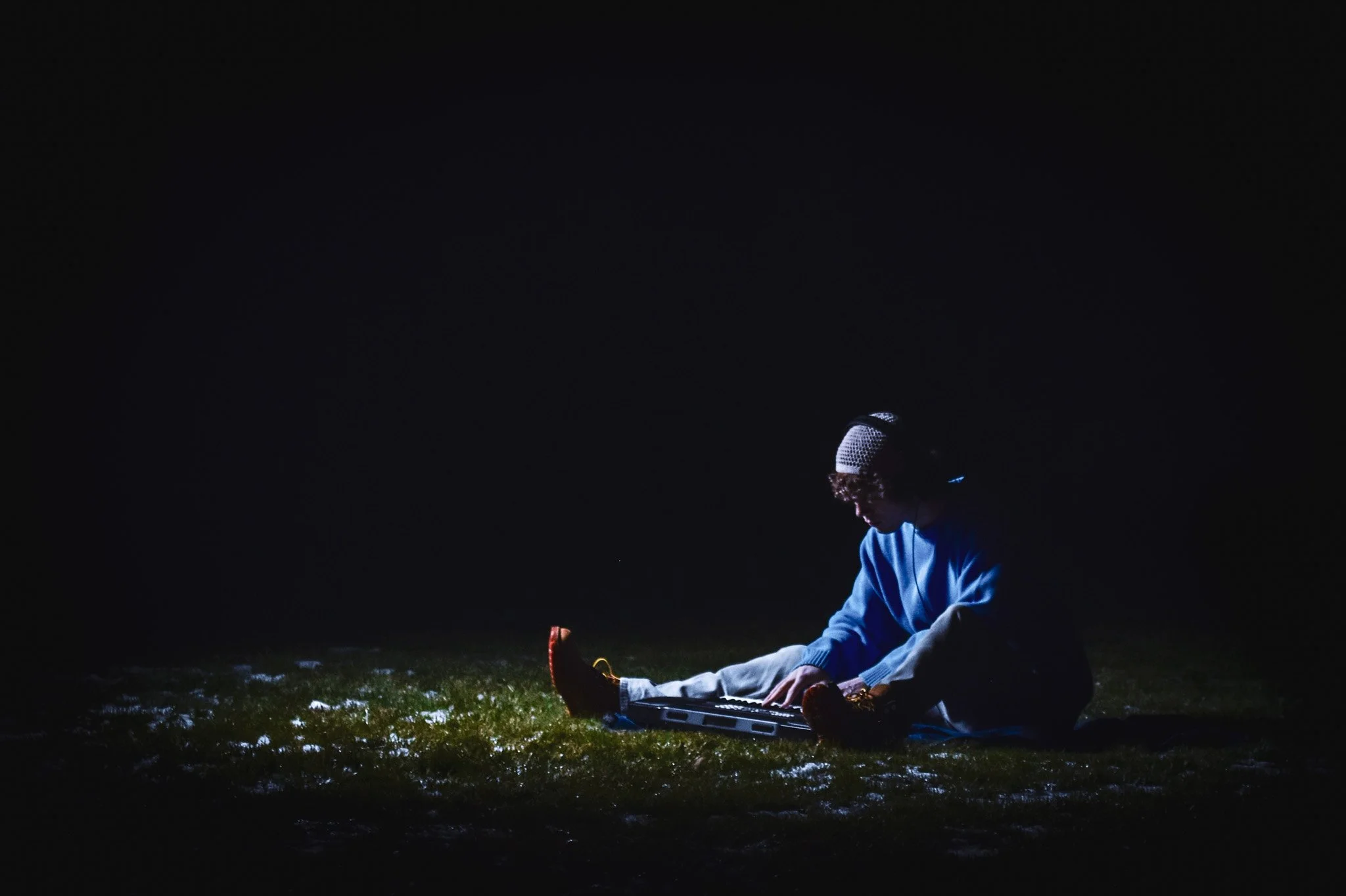 A person wearing a beanie and blue sweatshirt sitting outdoors at night, illuminated by a small light source, with a microphone nearby.