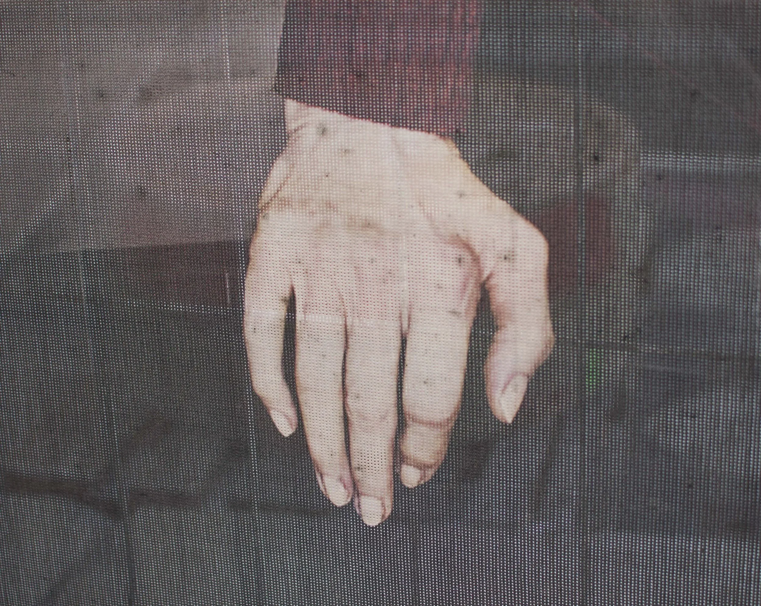 Die Farbfotografie einer Hand, offensichtlich von einer ältere Dame. Die Handfotografie ist abgebildet auf einer transparenten Planen an einem Bauzaun. Dahinter zeichnen sich Baumaterialien ab. Rostrote Farbanmutung.