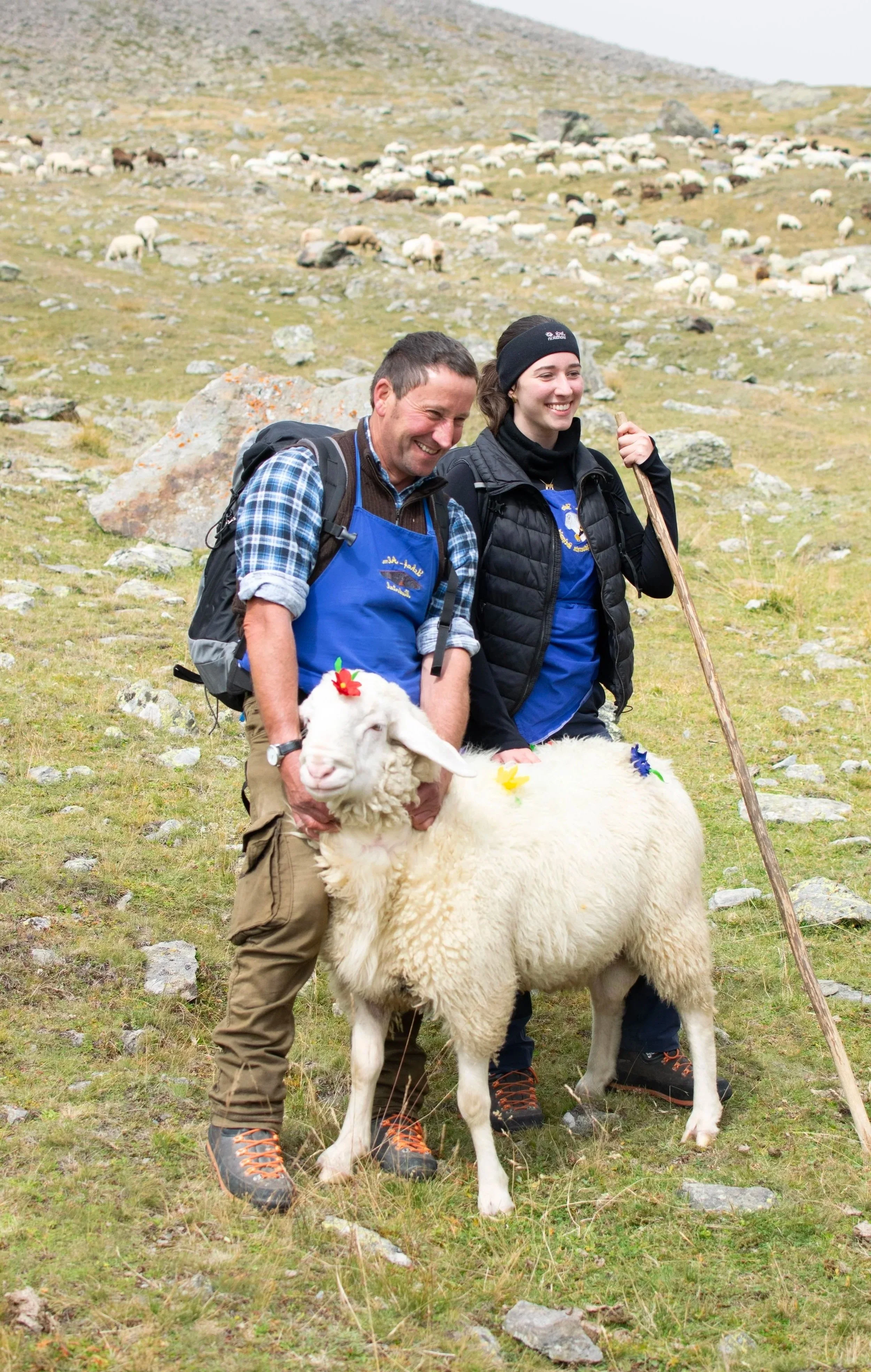 Vater und Tochter posieren stolz lachend mit einem geschmückten Schaf in einer bergigen, grasbewachsenen Landschaft. Hm Hintergrund eine Herde Schafe.