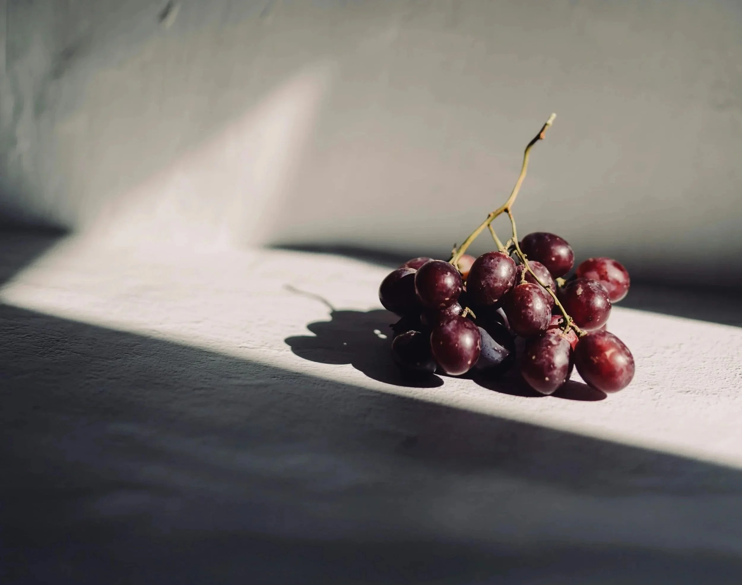 Eine Traube roter Trauben liegt auf einer weißen Oberfläche. Das Licht fällt von links oben und erzeugt Schatten nach rechts.