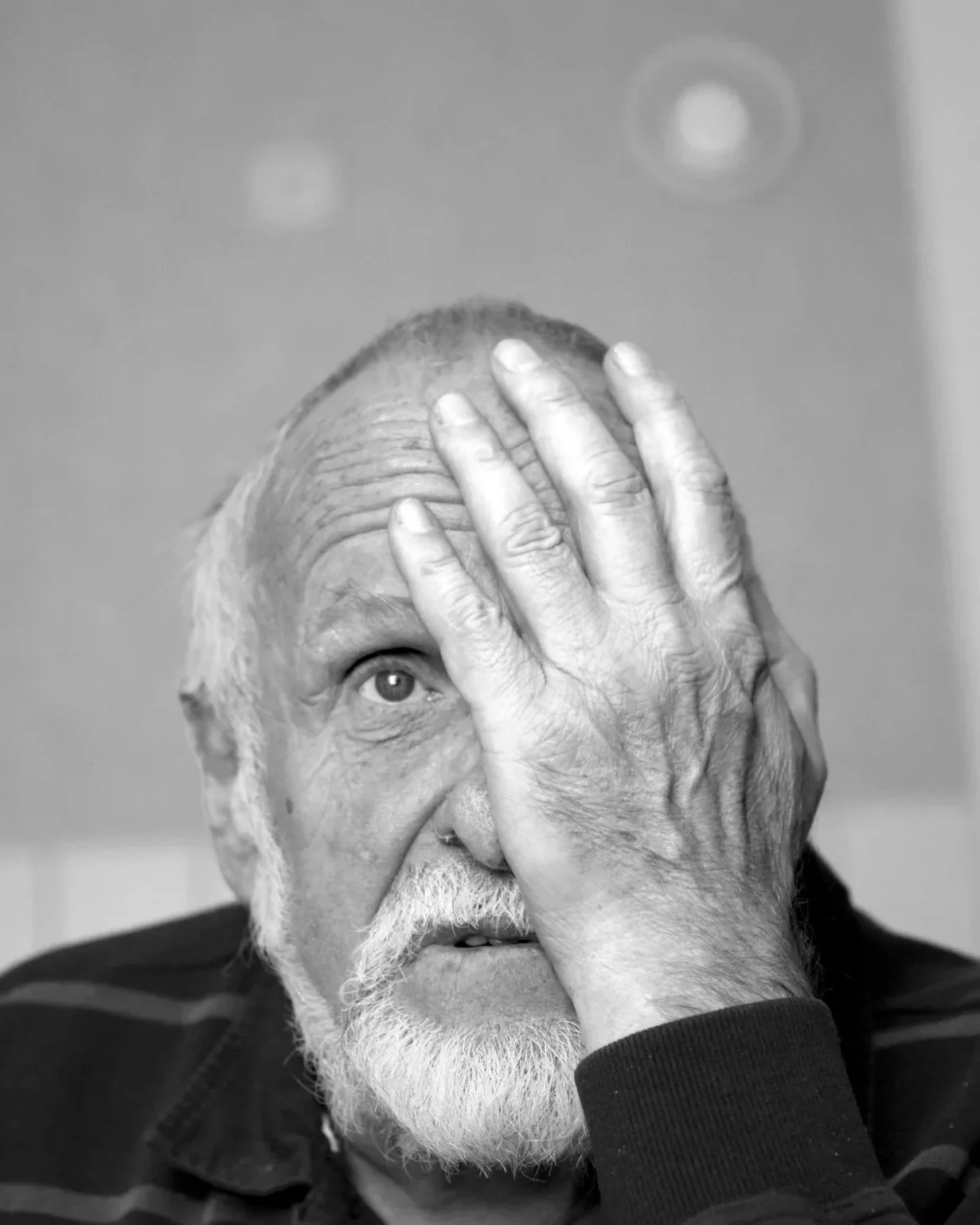 Schwarz-weiß-Fotografie eines älteren Mannes mit weißem Bart. Ein Auge hält er mit seiner Hand bedeckt. Der Mann schaut konsterniert. Hinter ihm zeichnet sich spartanisch eine Küchenwand ab.