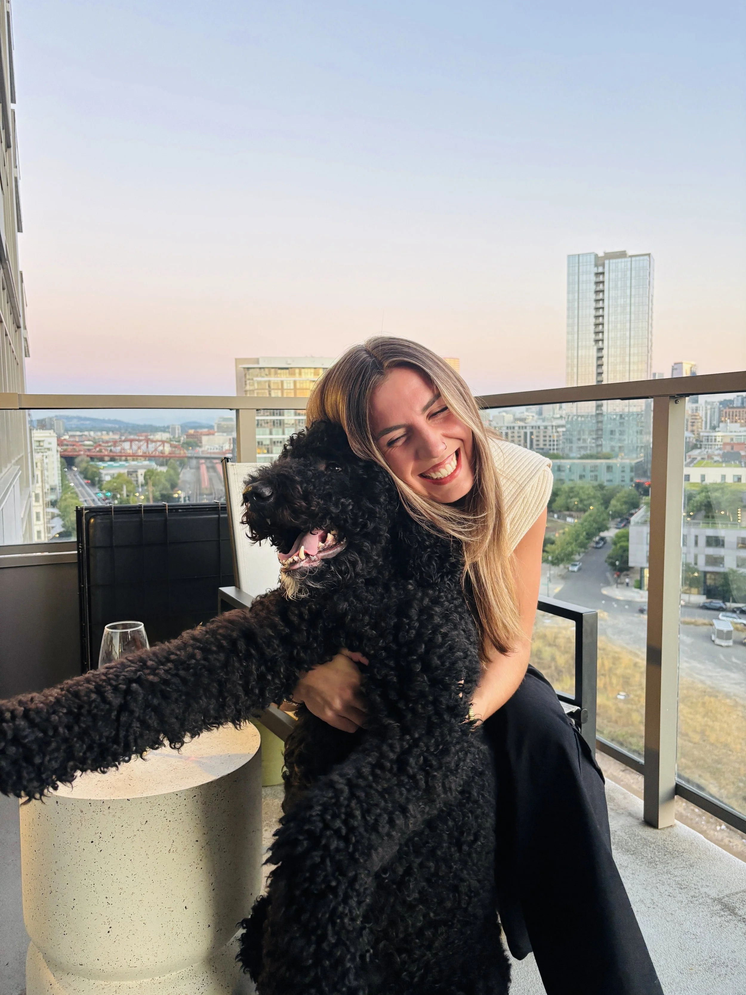 A woman with long blonde hair smiling and hugging a large black curly-haired dog on a balcony overlooking a cityscape at sunset.