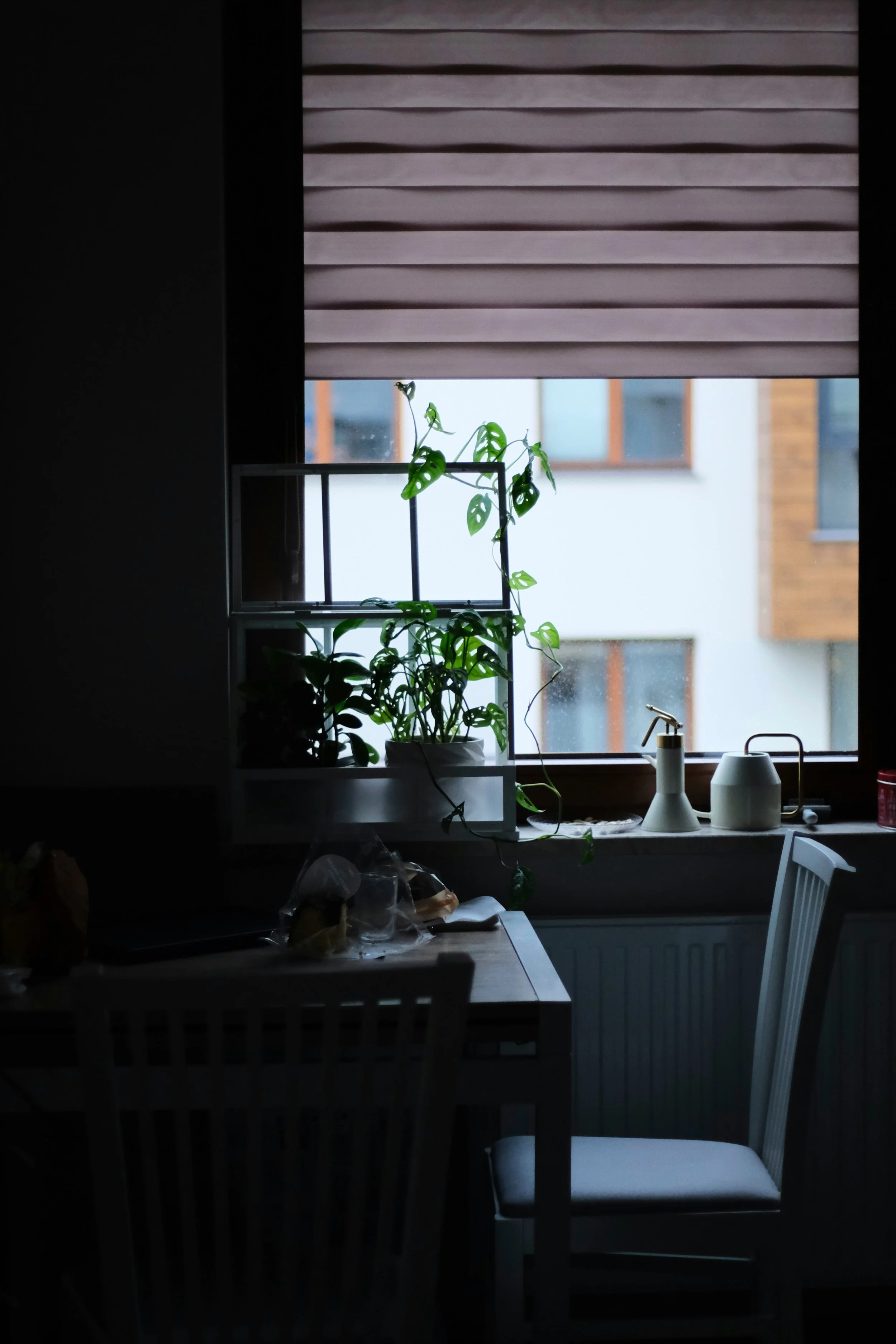 A room with a window partially covered by a rolled-up shade, and a small potted plant on the windowsill. The view outside the window shows neighboring building walls and windows. Inside, a table has some items, including a bread loaf in plastic wrap, and there are chairs around the table.