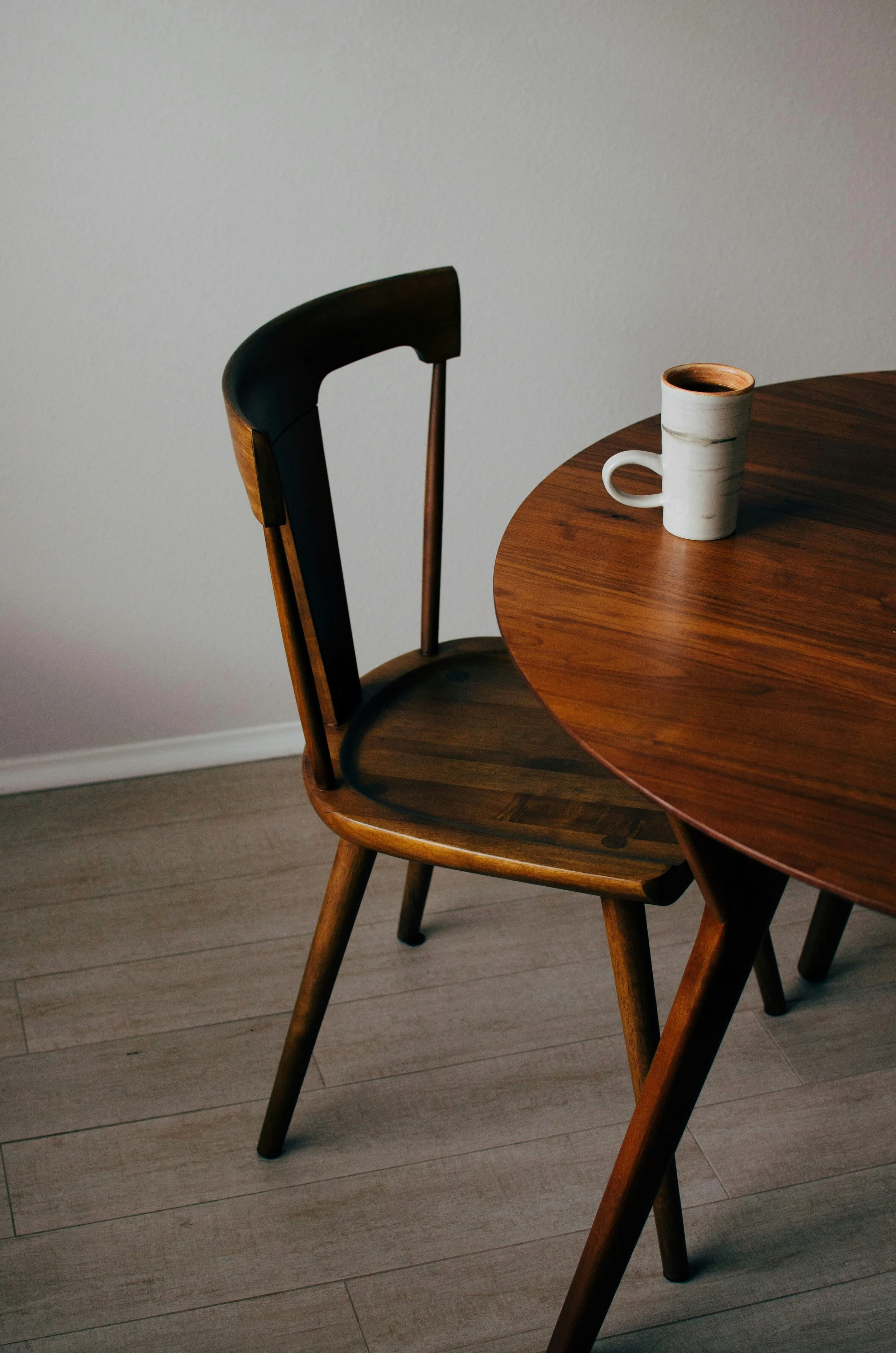 Close-up of a wooden dining table with a white and gray ceramic mug on it, against a plain light-colored wall.