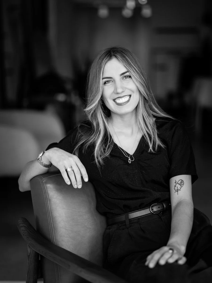 Black and white photo of a woman sitting on a chair, smiling at the camera. She has shoulder-length wavy hair and is wearing a dark shirt, jewelry, and has a tattoo on her left arm.