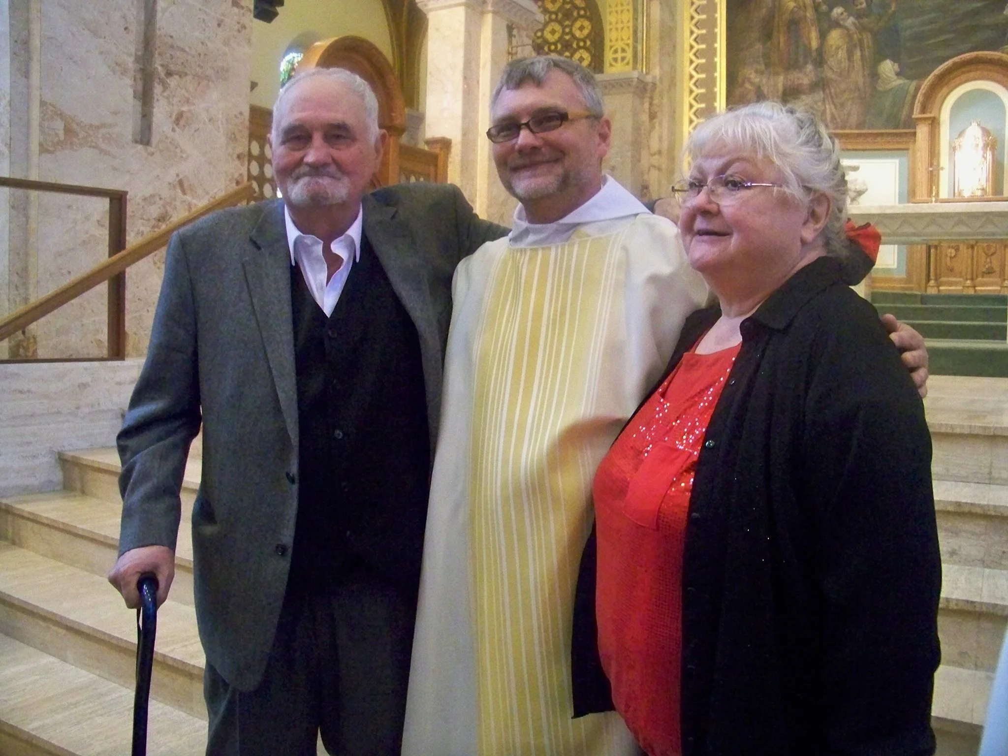 Three people in a church, standing on the stairs. Left is an older man with a cane, wearing a grey blazer. In the middle, a man wearing glasses and a white robe with yellow stripes. On the right, an older woman with white hair and glasses, wearing a black jacket and a red dress.