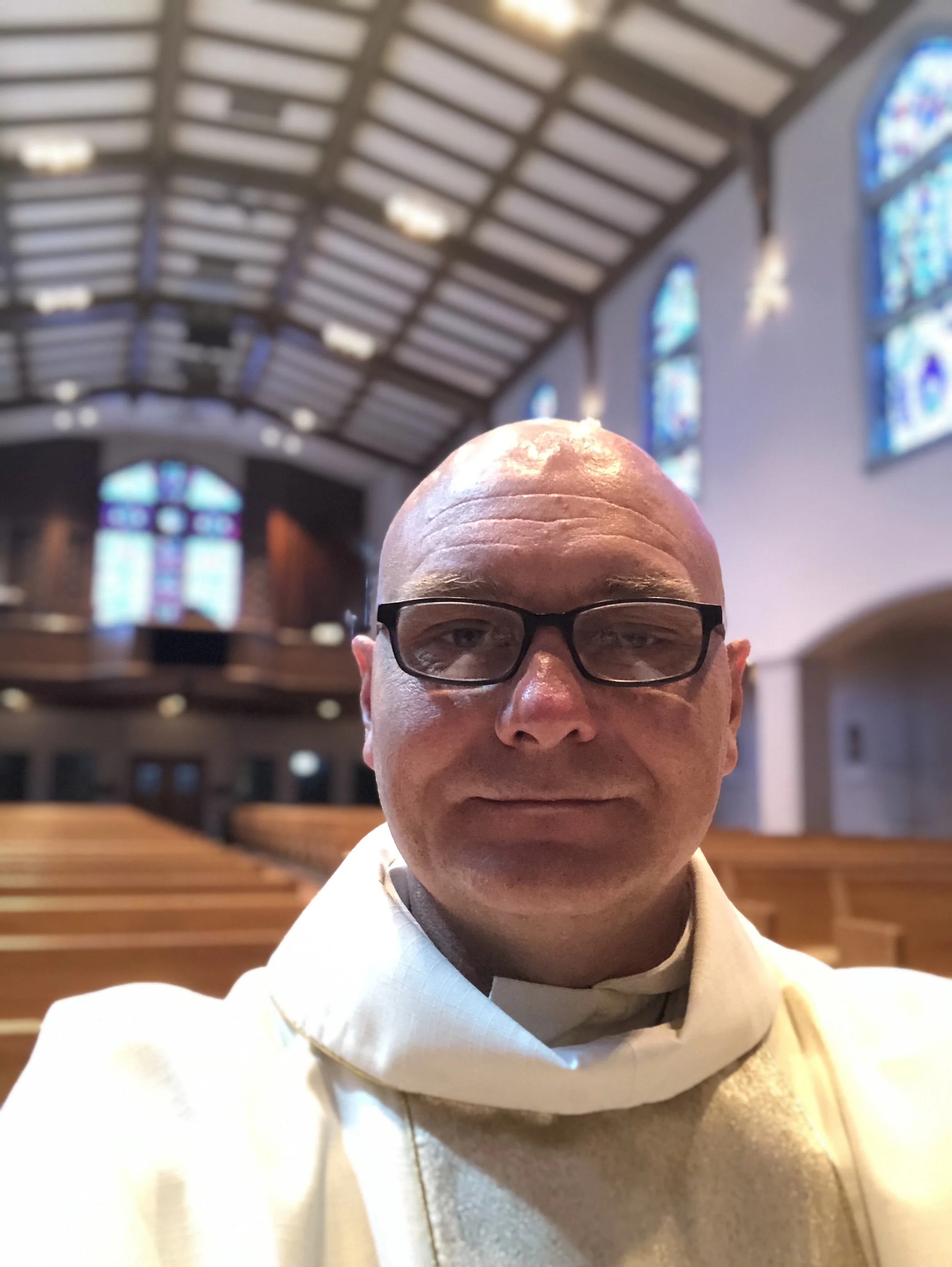 A man with glasses taking a selfie inside a church with wooden pews and stained-glass windows.