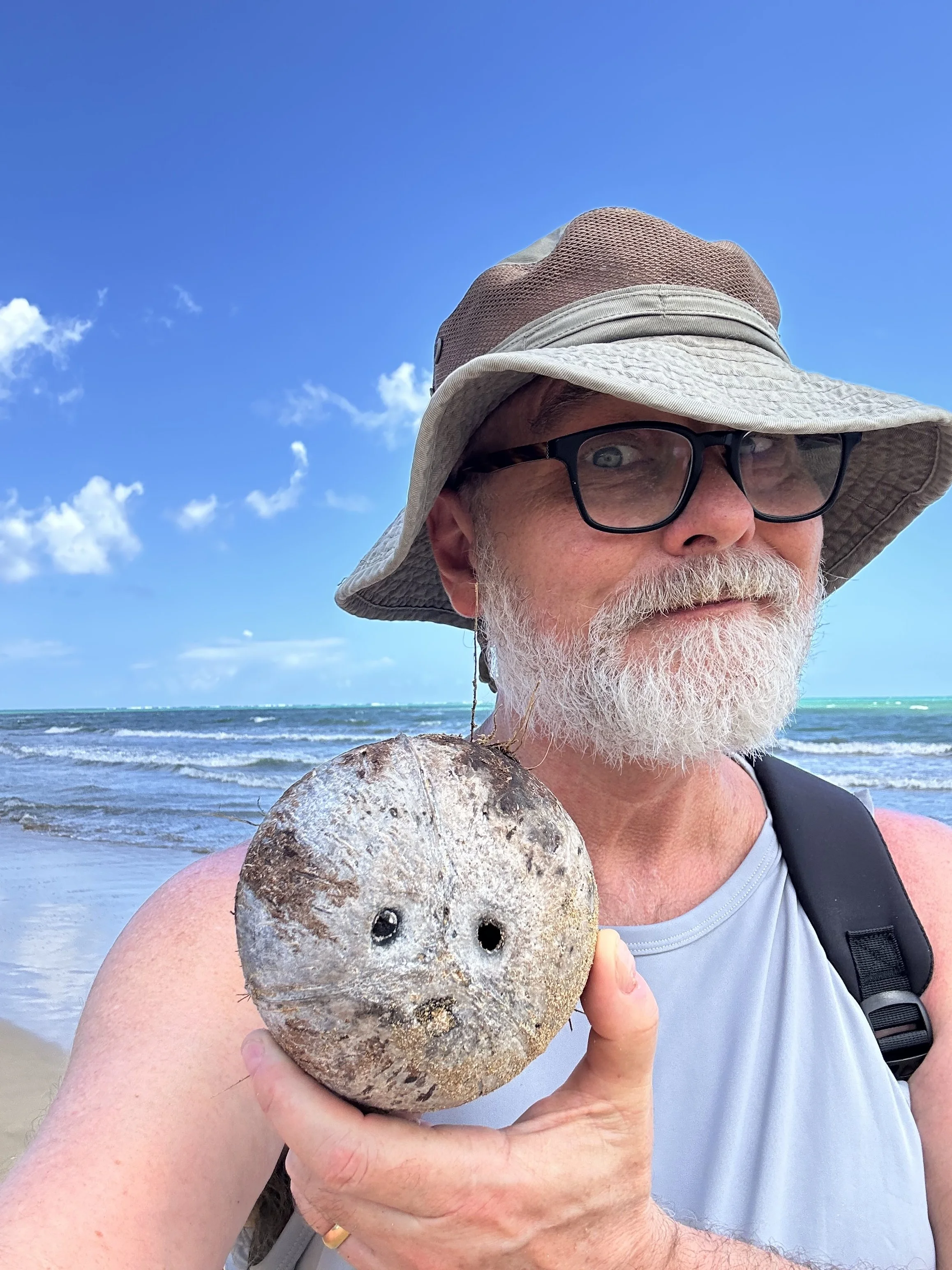 A man with glasses and a hat is holding a large, weathered seashell near his shoulder at the beach, with the ocean and a blue sky with some clouds in the background.