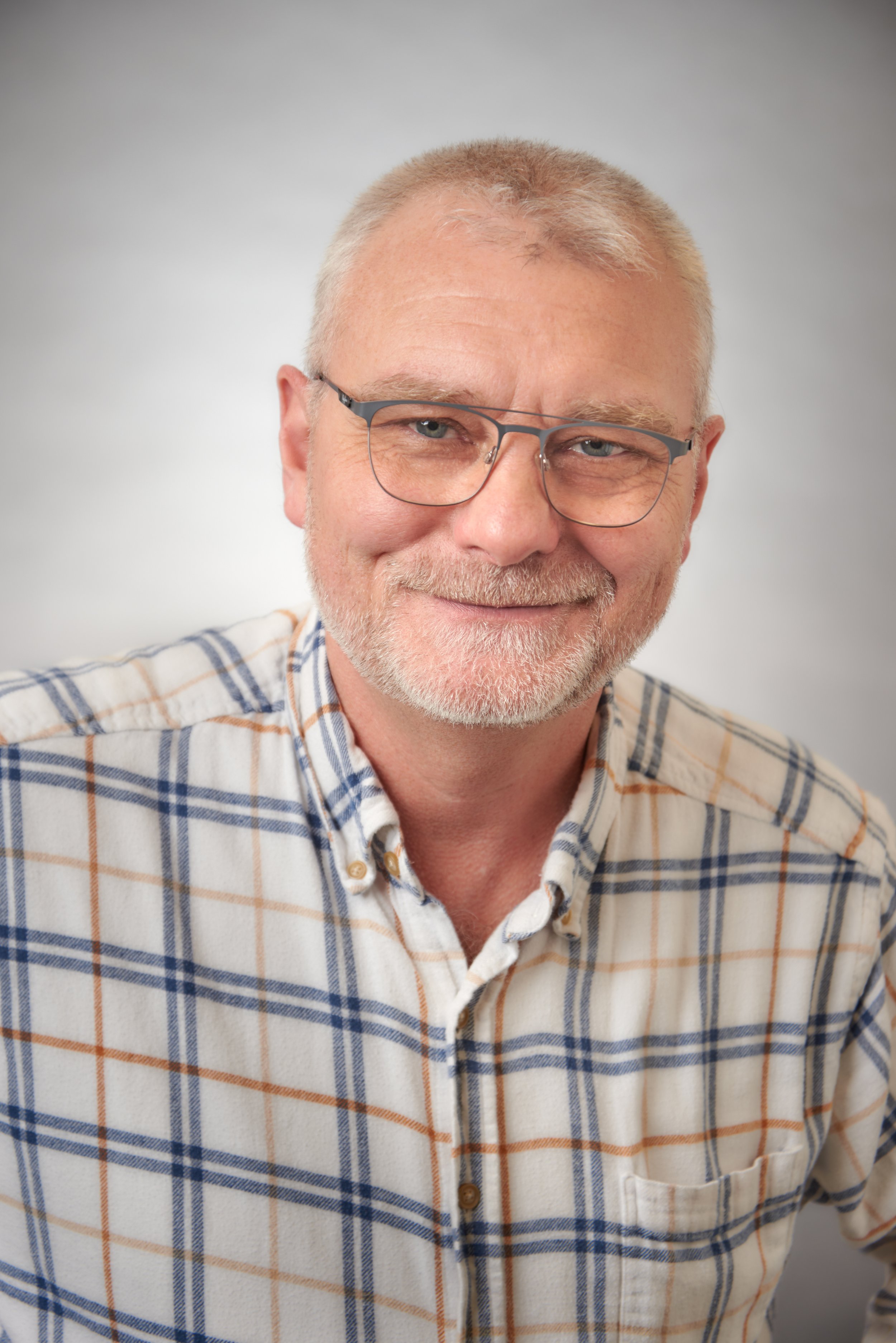 A middle-aged man with short gray hair, glasses, and a beard smiling, wearing a plaid shirt.