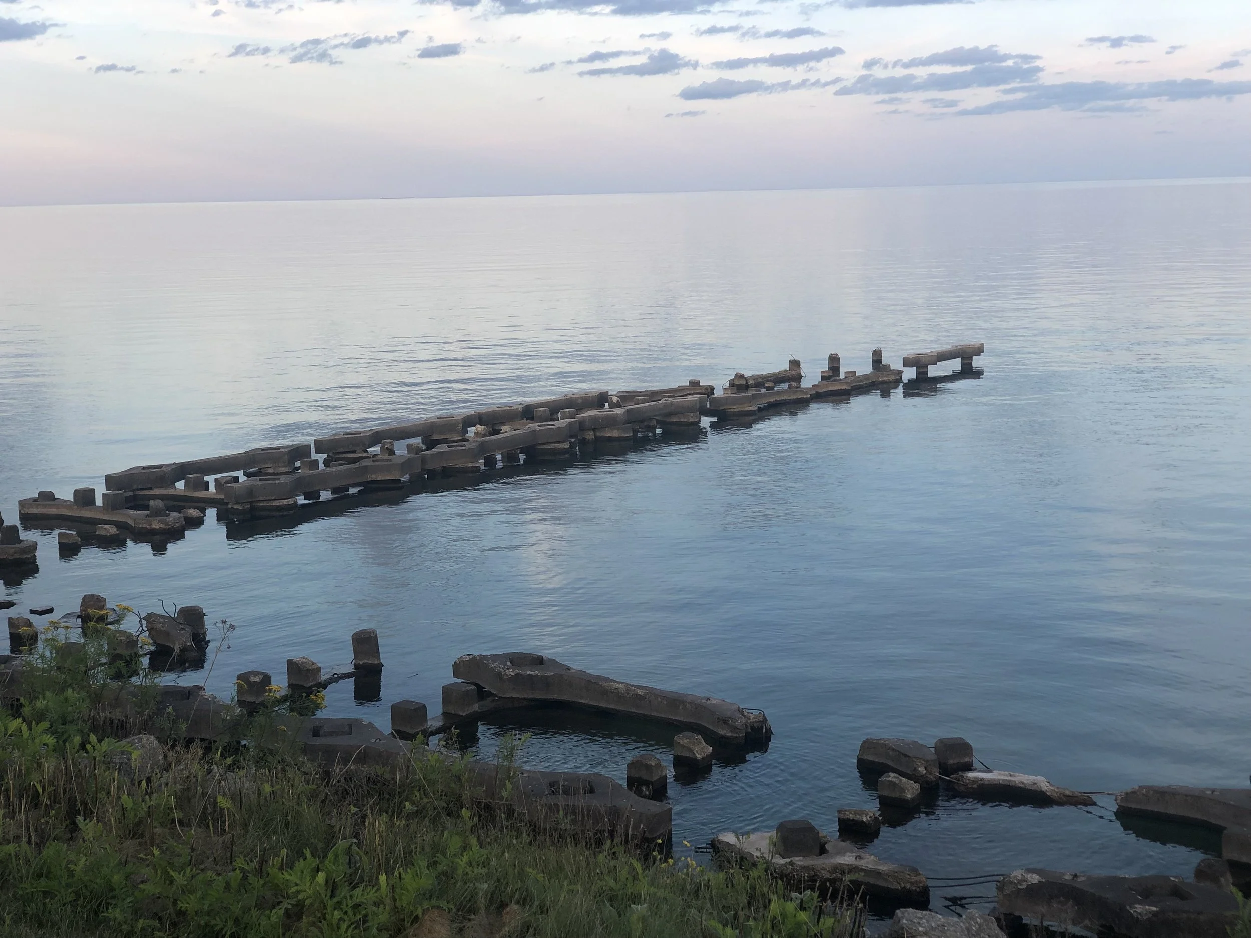 A calm body of water with a wooden pier extending into it, surrounded by grass and rocks, under a cloudy sky.