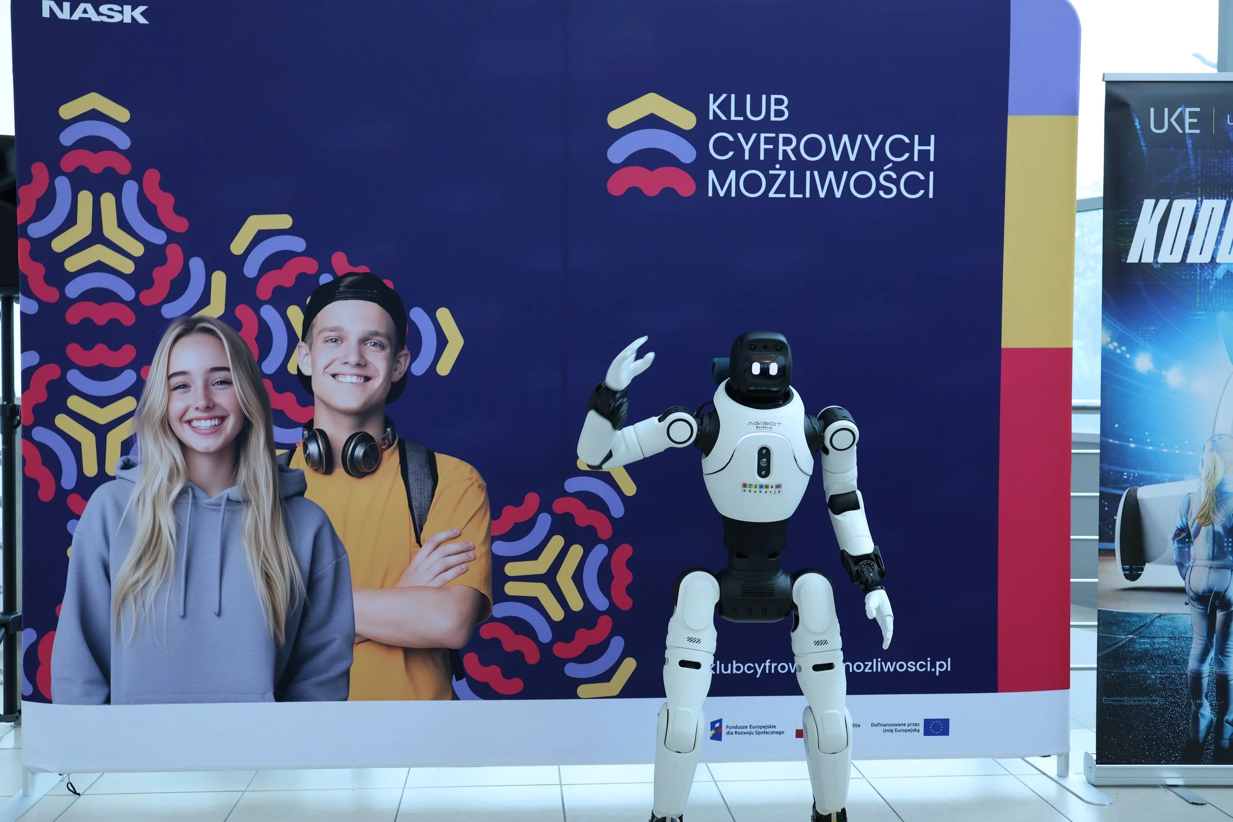 Humanoid Xawery z Embodied AI Lab macha na powitanie przed ścianką Klubu Cyfrowych Możliwości NASK na Dniu Cyfrowych Możliwości we Wrocławiu.