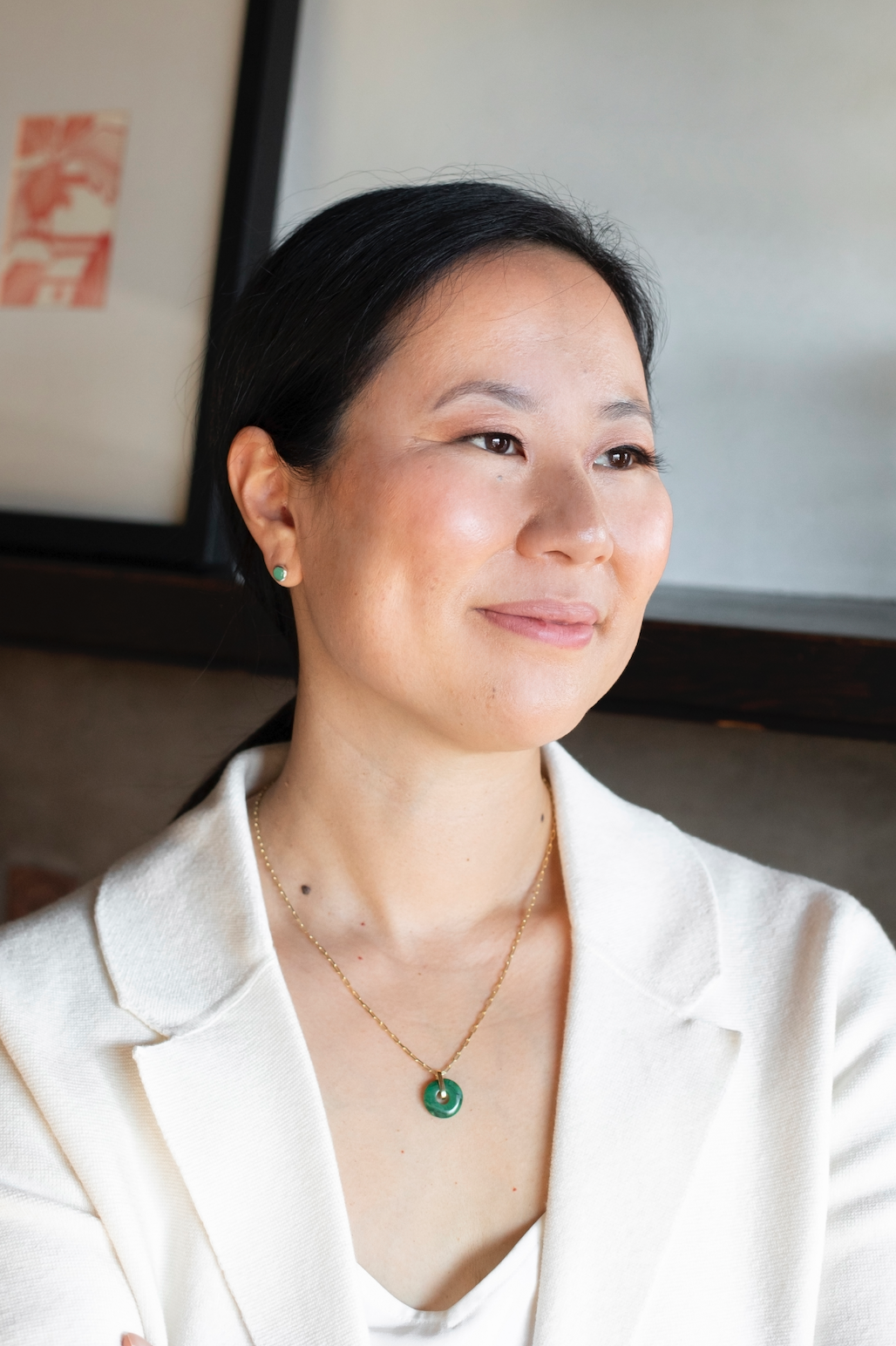 A woman with dark hair wearing a white blazer, green jewelry, and a necklace, looking to the right and smiling softly.