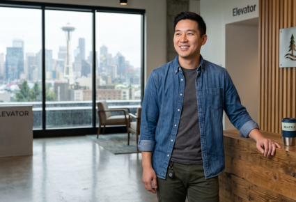 A young man with a coffee cup standing in a modern office lobby with a skyline view of Seattle in the background.