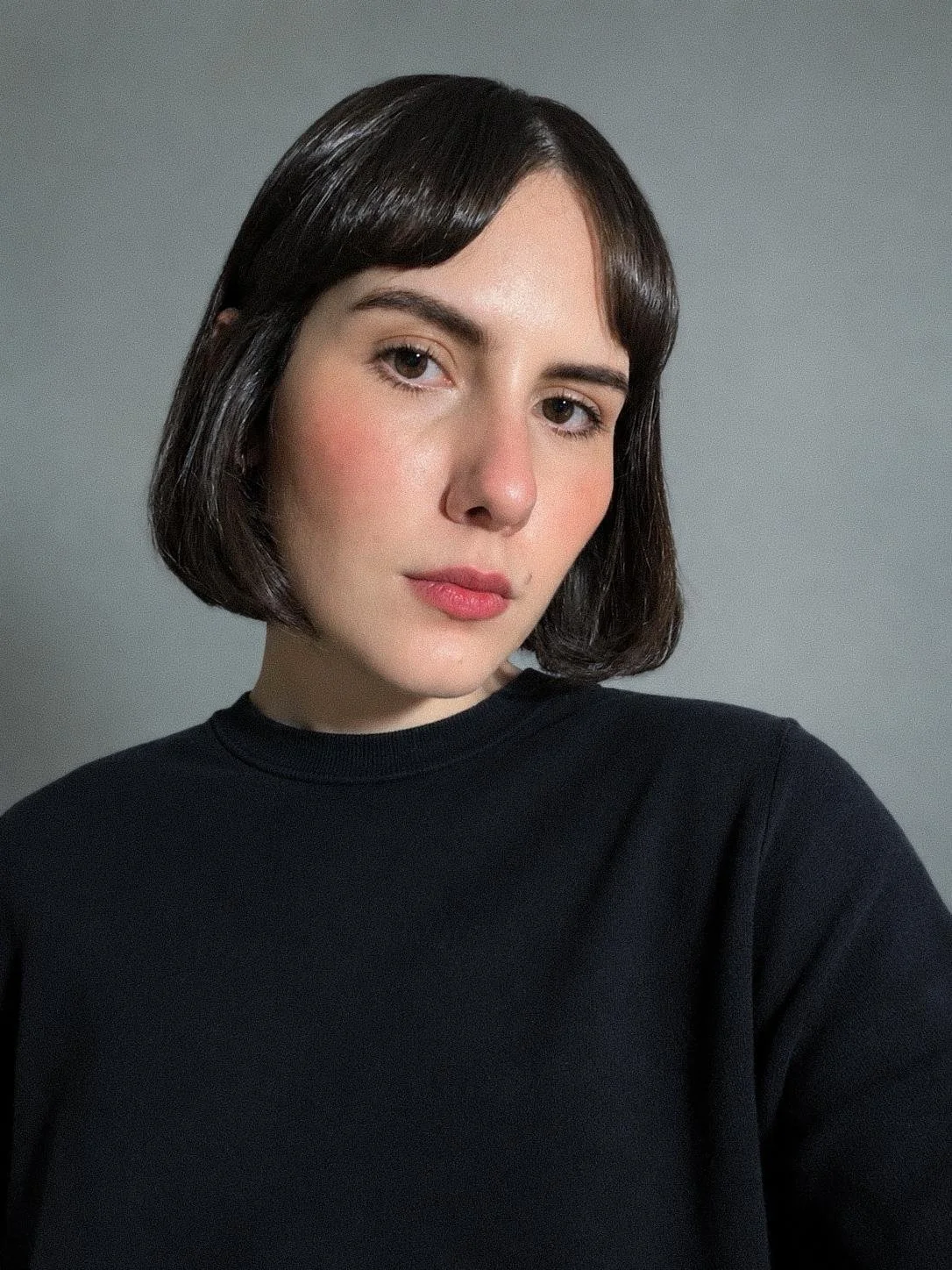 A young woman with short dark hair and fair skin wearing a black shirt, looking at the camera against a plain gray background.