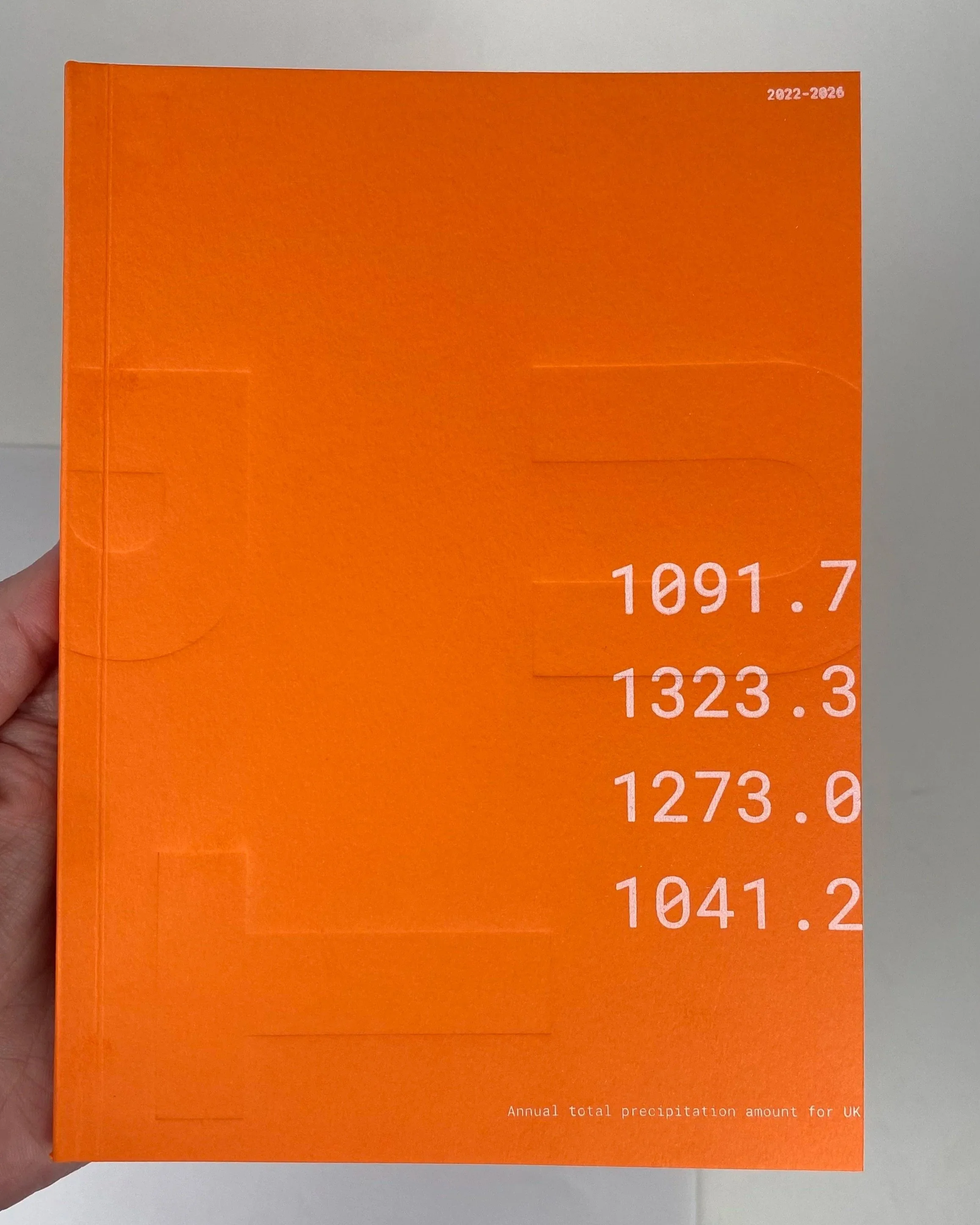 Orange booklet with white text on the cover showing annual total precipitation amounts for the UK from 2022 to 2026, with specific figures listed.