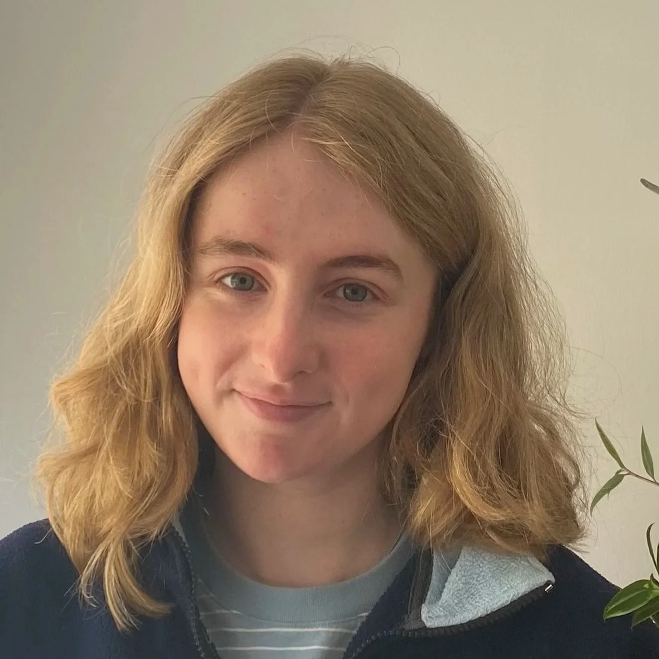A young woman with wavy, shoulder-length blonde hair, blue eyes, and light skin, smiling slightly. She is wearing a dark jacket over a gray shirt with white stripes and standing indoors beside a plant with green leaves.