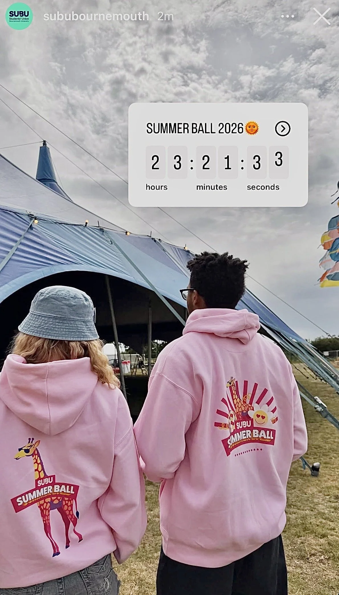 Two people from behind, wearing pink hoodies with a retro summer event logo featuring a giraffe, at an outdoor fairground. The logo reads 'SUBU Summer Ball' with a giraffe and heart emoji. The sky is cloudy, with a tent and some equipment visible in the background.
