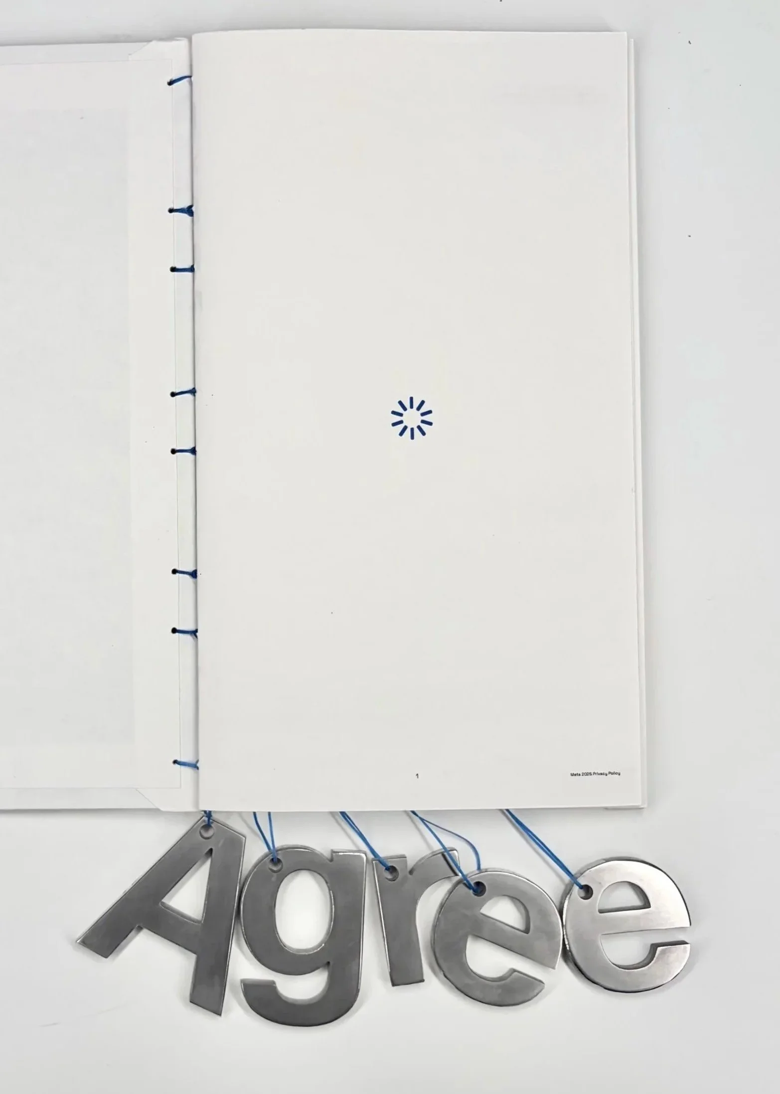 A photo of a white folder or binder with a blue spiral binding, on which a silver decorative sign displaying the word 'Agree' is hanging using blue string. The photo appears to be taken from above.