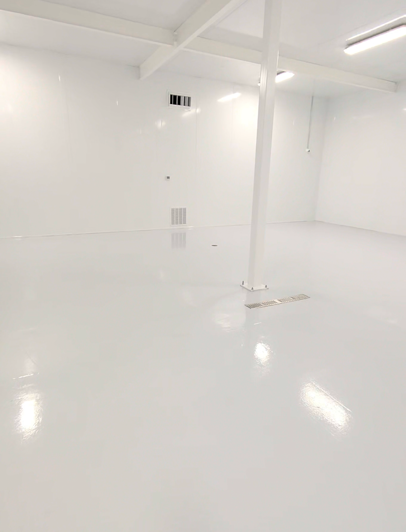 An empty, clean, white industrial or storage room with a shiny floor, white walls, and ceiling. There are vents and ducts on the wall, and a drain on the floor.