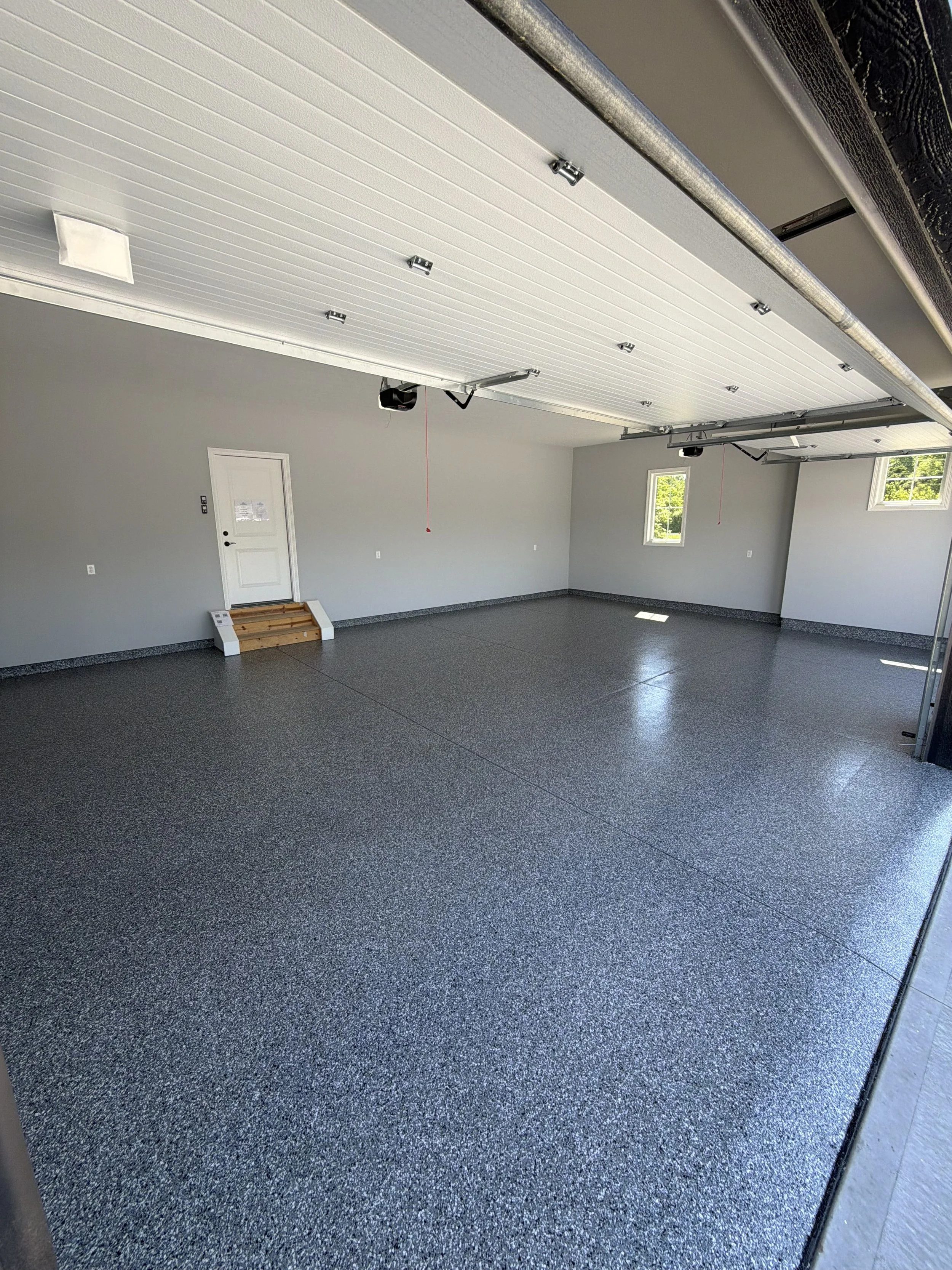Interior view of a garage with gray walls, a speckled gray floor, windows, and a partially open garage door.