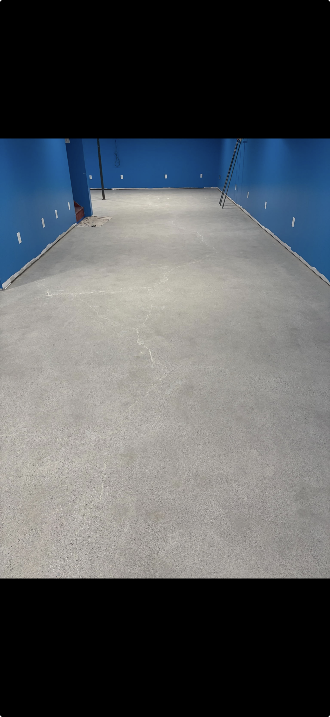 Empty room with blue walls, concrete floor that's grinded and had crack repair, and some construction materials and tools leaning against the wall.