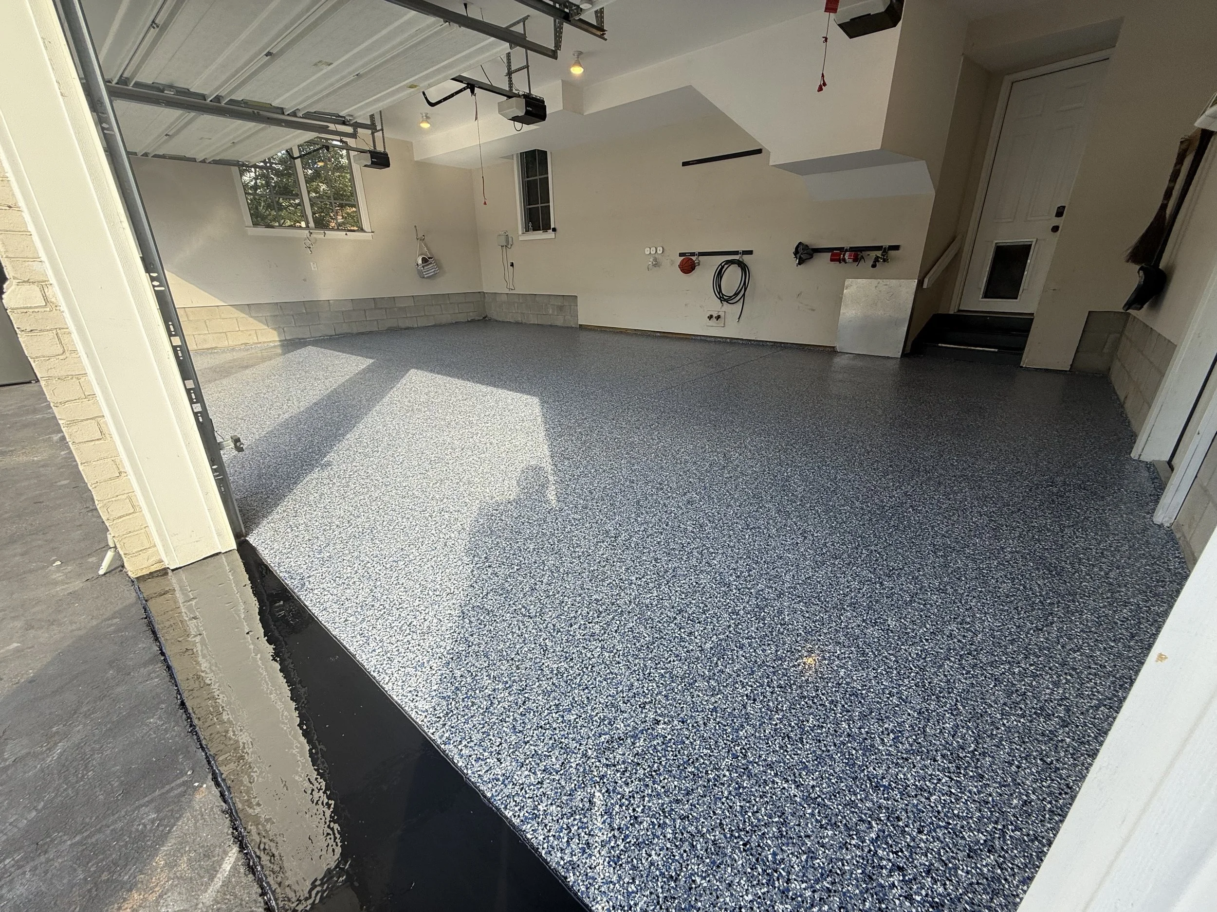 View of a 3 car garage with flake epoxy floor with the color being orbit, a white ceiling with recessed lighting, and a partially open garage door. There are windows on the back wall and some tools, hoses, and wall-mounted fire extinguishers visible.