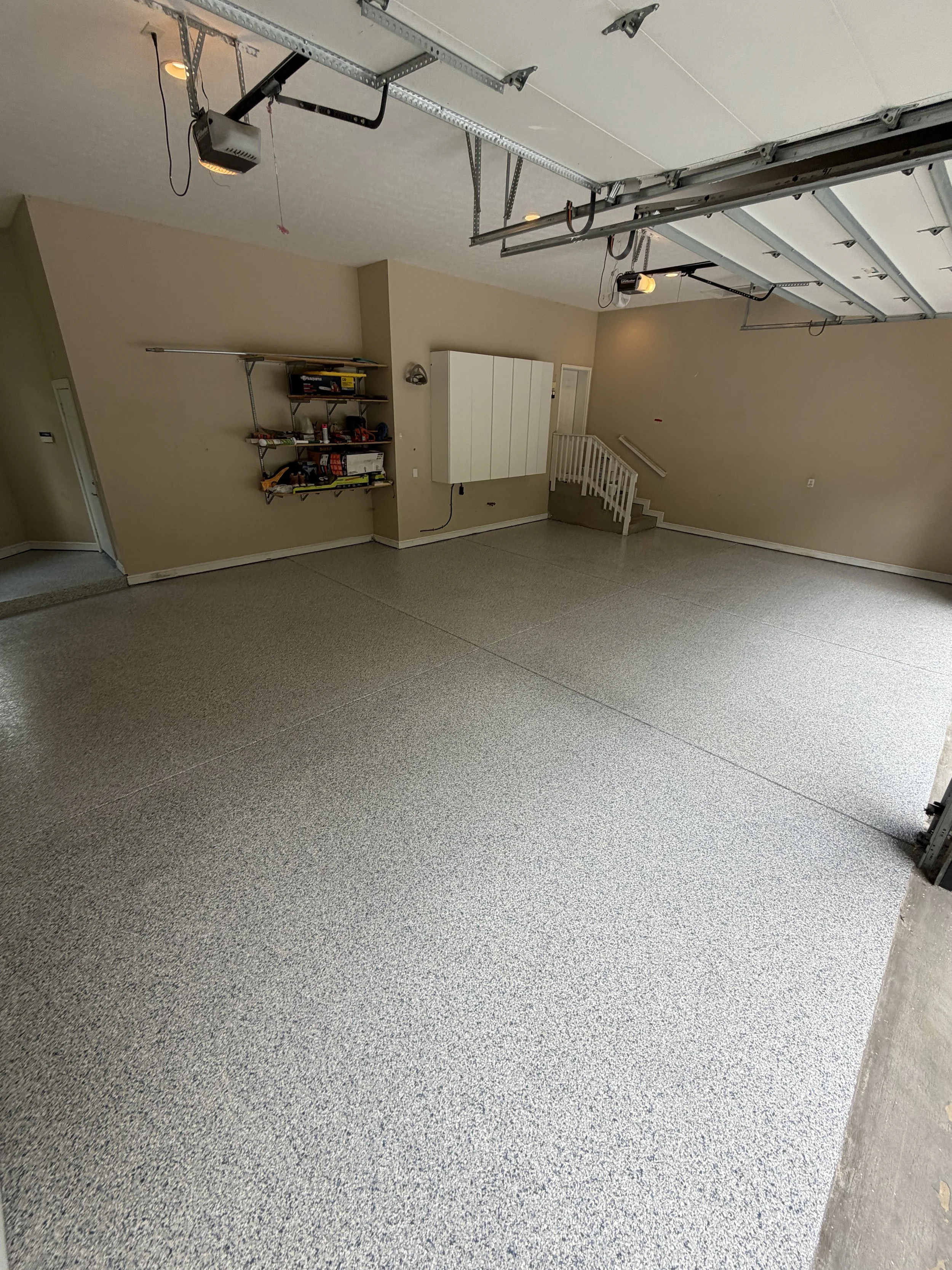 A newly finished garage with a flake epoxy floor, beige walls, and a partially open garage door. There are tools on a wall shelf and some electrical and garage door openers installed.