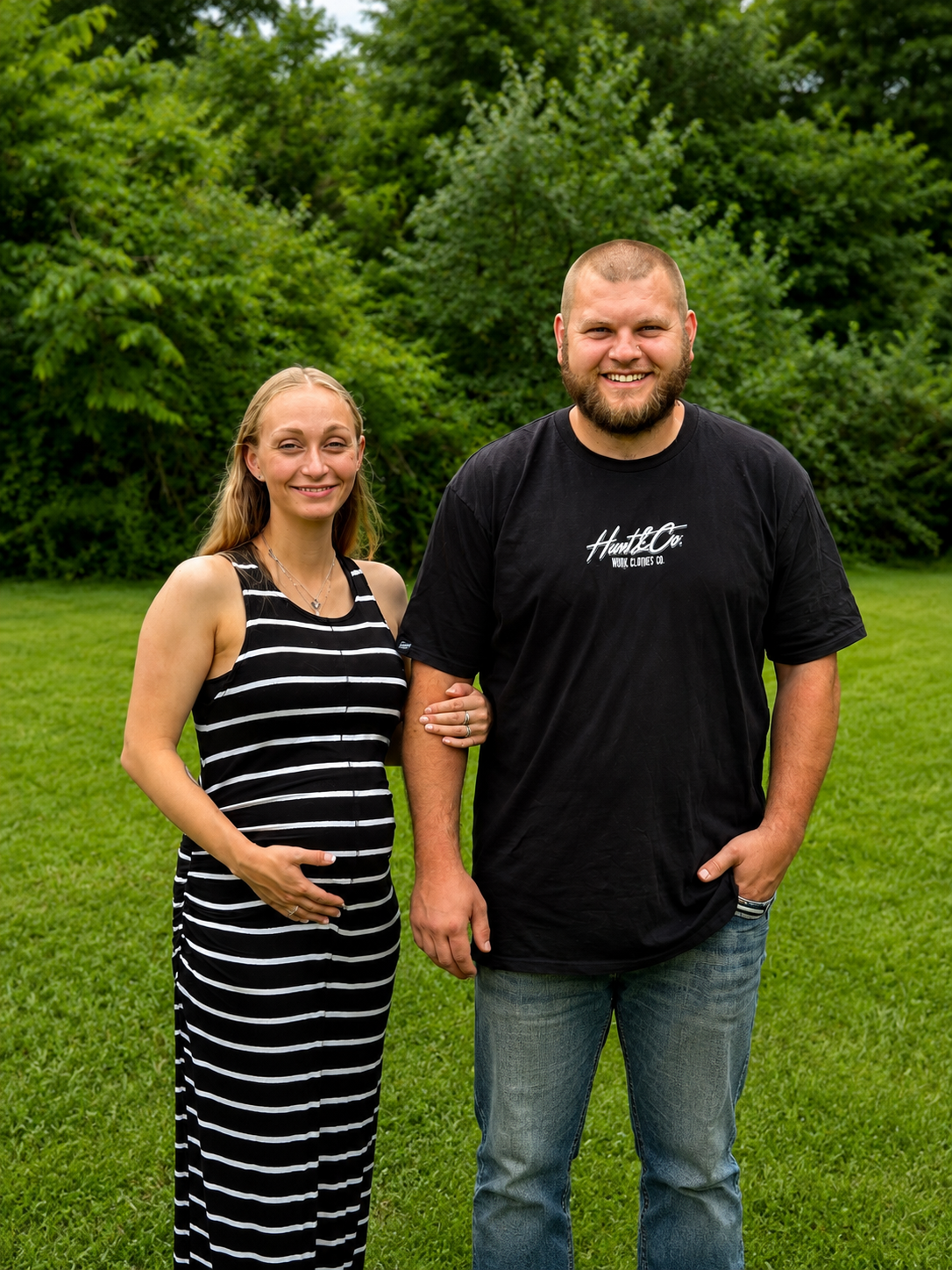 A smiling woman with long blonde hair in a black and white striped dress standing next to a smiling man with short hair and beard, wearing a black T-shirt and jeans, outdoors with green trees and grass in the background.