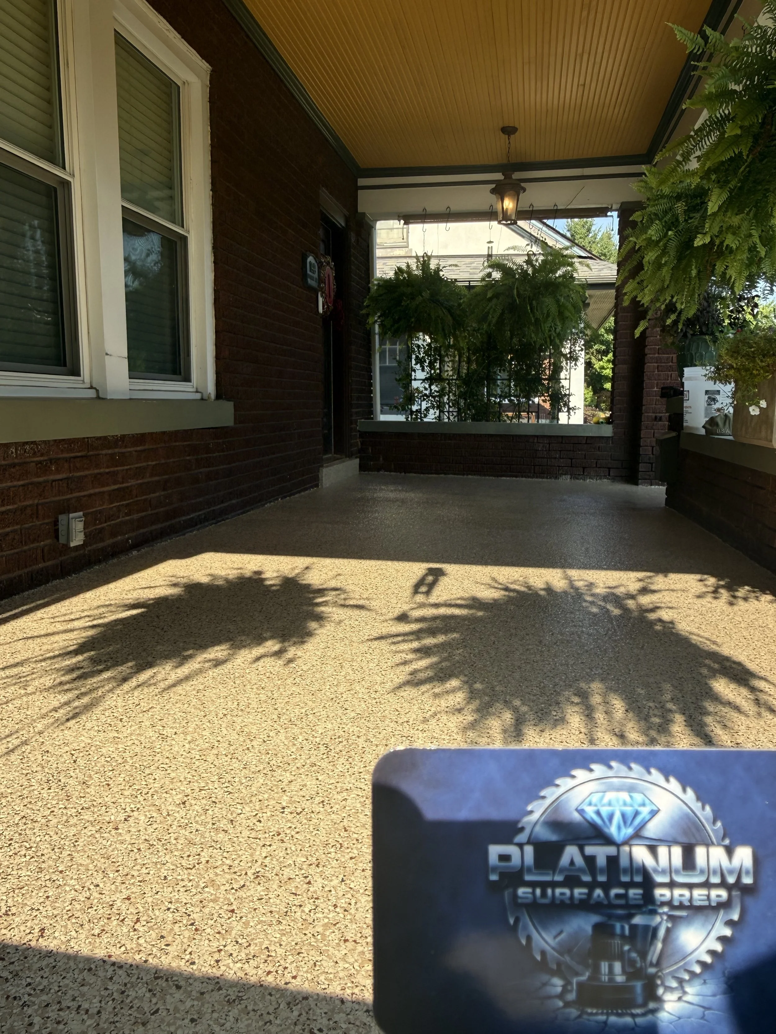 Font porch of a house with brick walls, a yellow wooden ceiling, hanging plants, and a hanging light fixture. Epoxy flake floor with the flake color being cliffs edge.