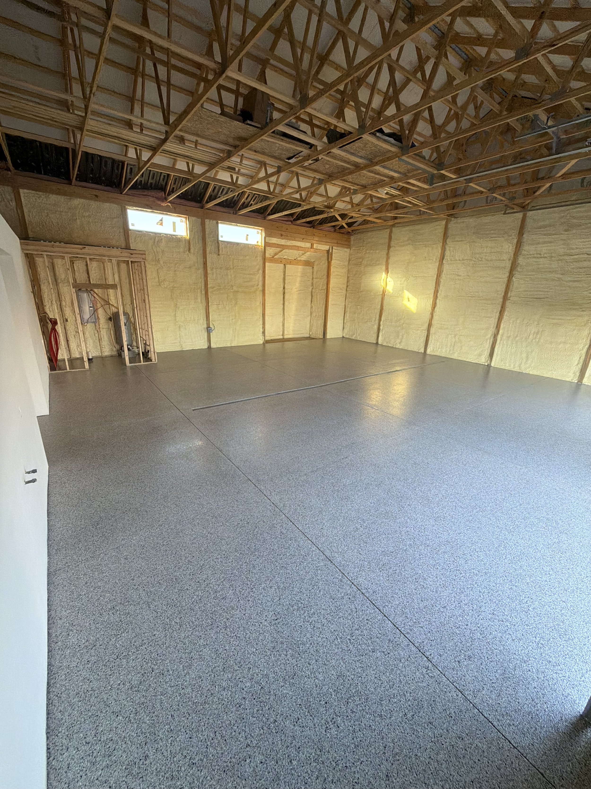 Room under construction with exposed wooden roof trusses, unfinished walls with insulation, and a finished gray speckled floor.