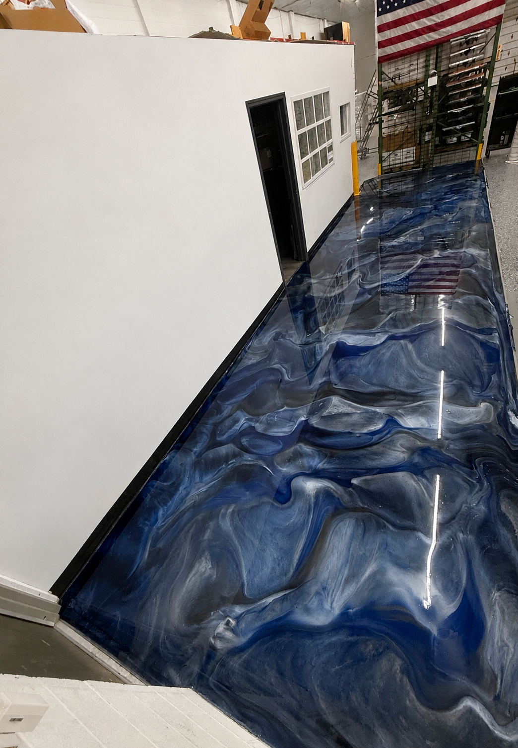 Indoor space with a glossy blue and white marbled epoxy floor, white partition wall, door with glass panels, and American flag hanging on the wall.