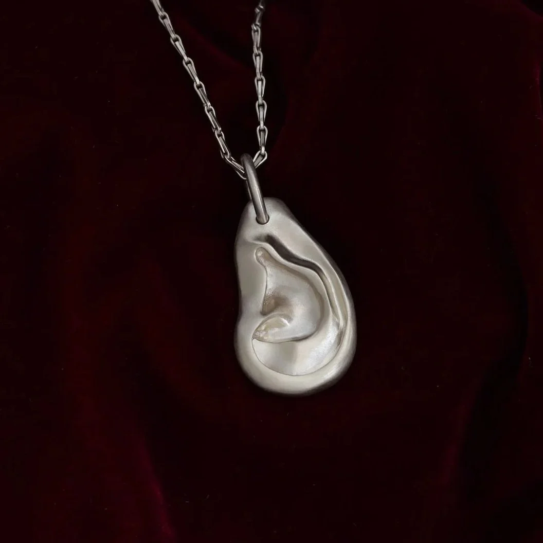 A silver necklace with a pendant shaped like a human ear, hanging against a dark background.