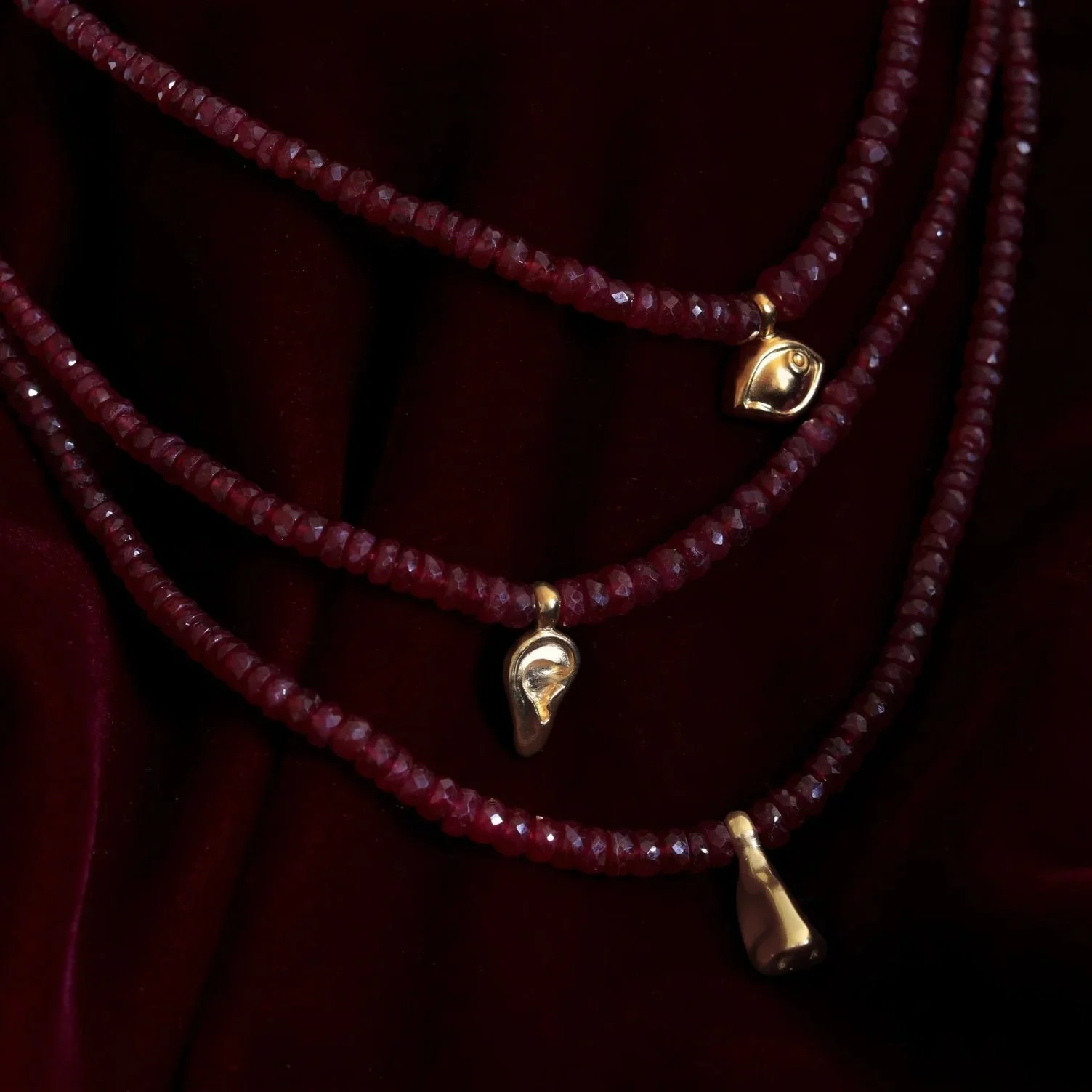 Red ruby necklace with charms -  an eye, an ear, and a nose charm, resting on a dark red velvet fabric.