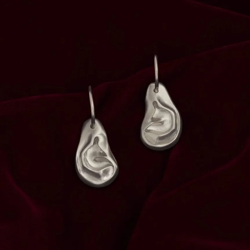 A pair of ear-shaped earrings dangling on hoops on a dark red, velvet background. 