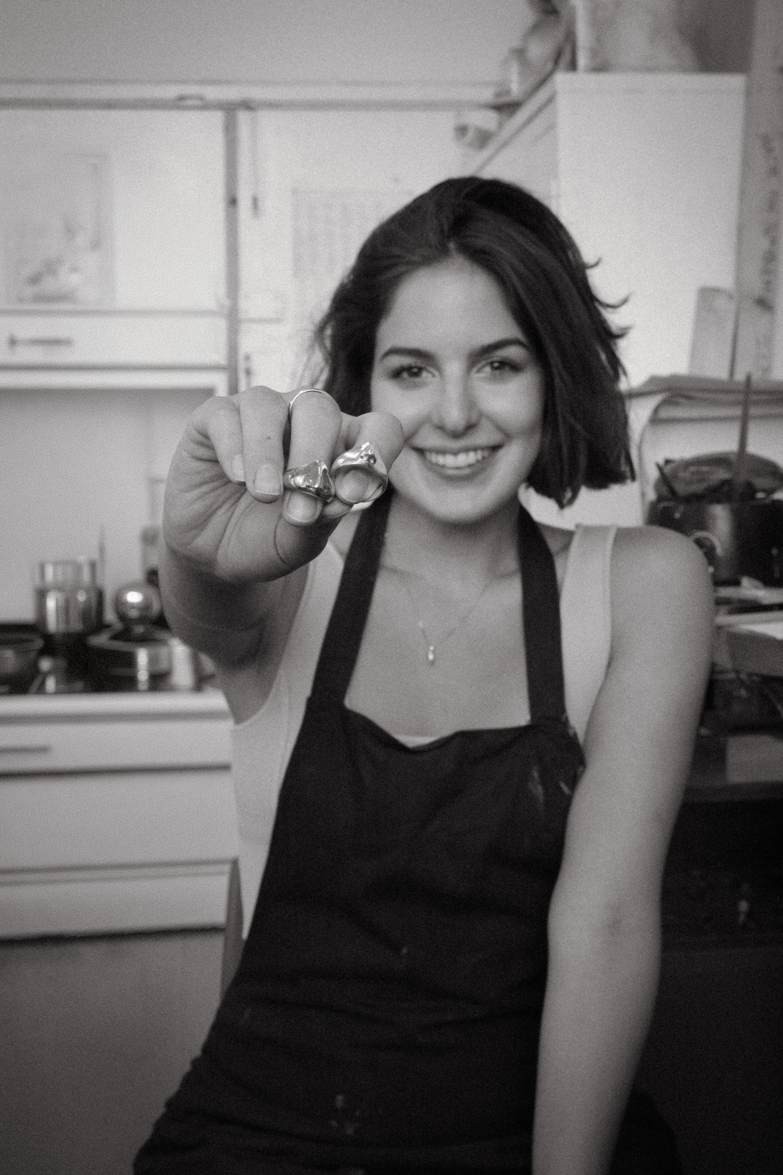 A woman with dark, shoulder-length hair smiling and showing rings on her fingers in jewellery studio, wearing a dark apron.