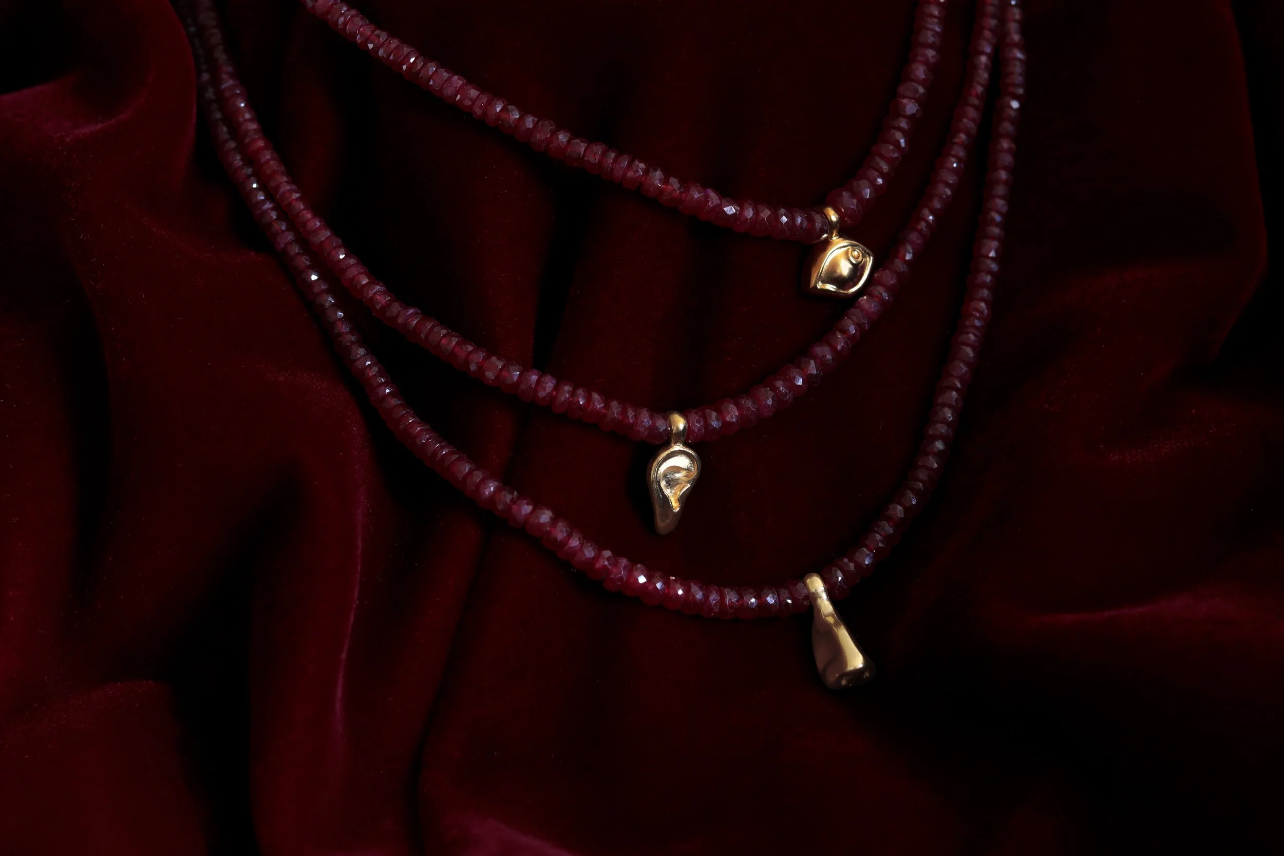 Red ruby beaded necklace with gold eye, nose, and ear pendants on deep red velvet fabric.