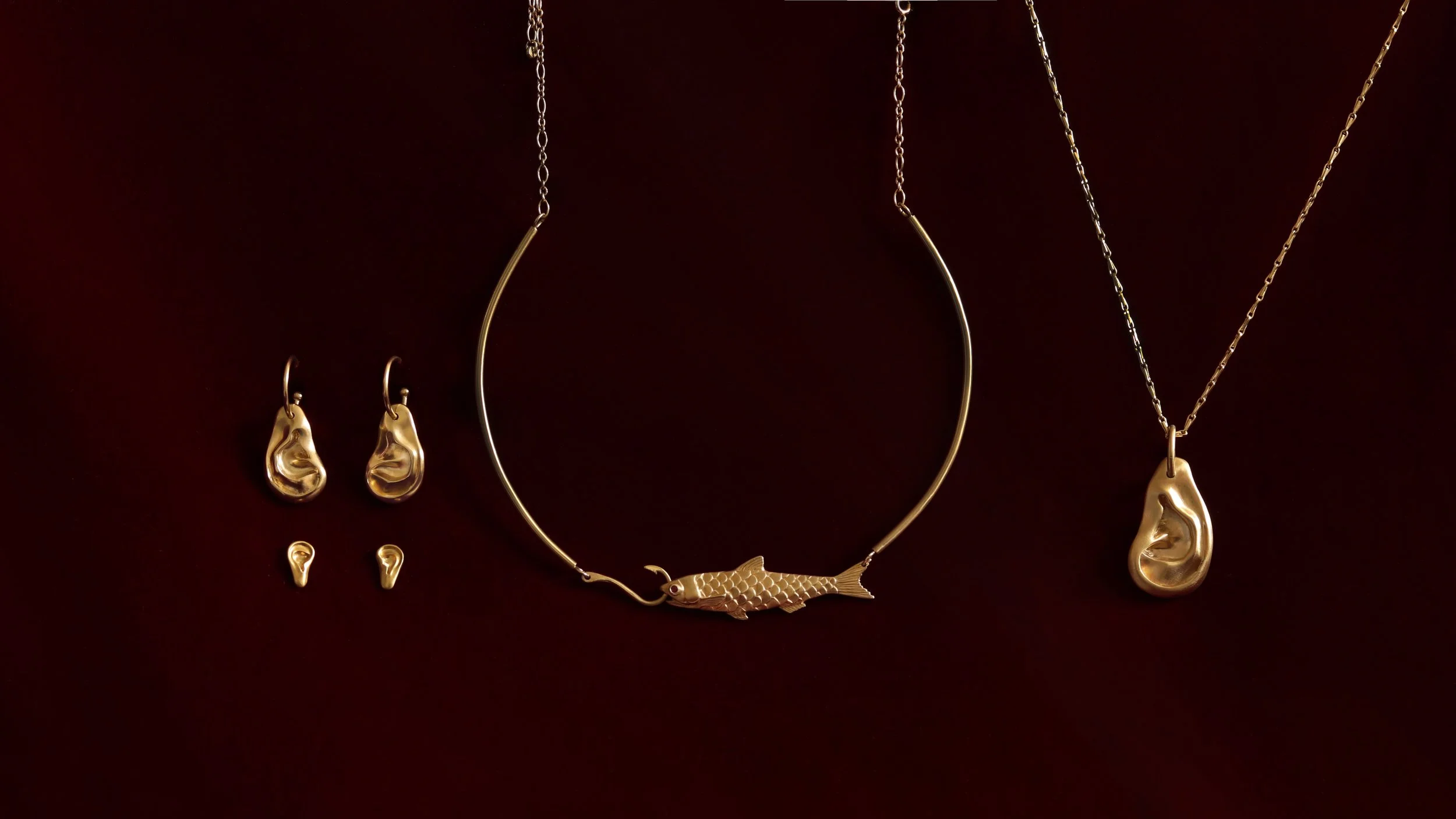Gold jewelry collection consisting of a fish necklace, ear-shaped earrings, and an ear-shaped necklace with a textured surface, all displayed against a dark background.