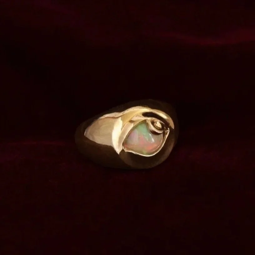A gold eye-shaped ring with a large, oval-shaped opal gemstone, set against a dark background.