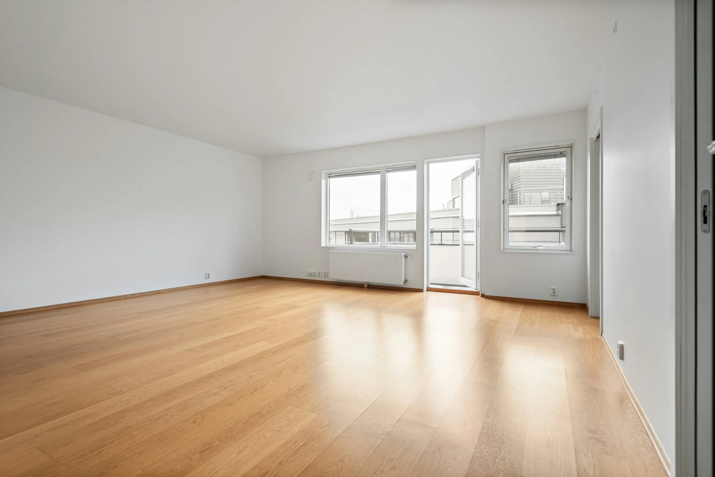 Empty room with white walls, wooden floor, large windows, and a balcony door.