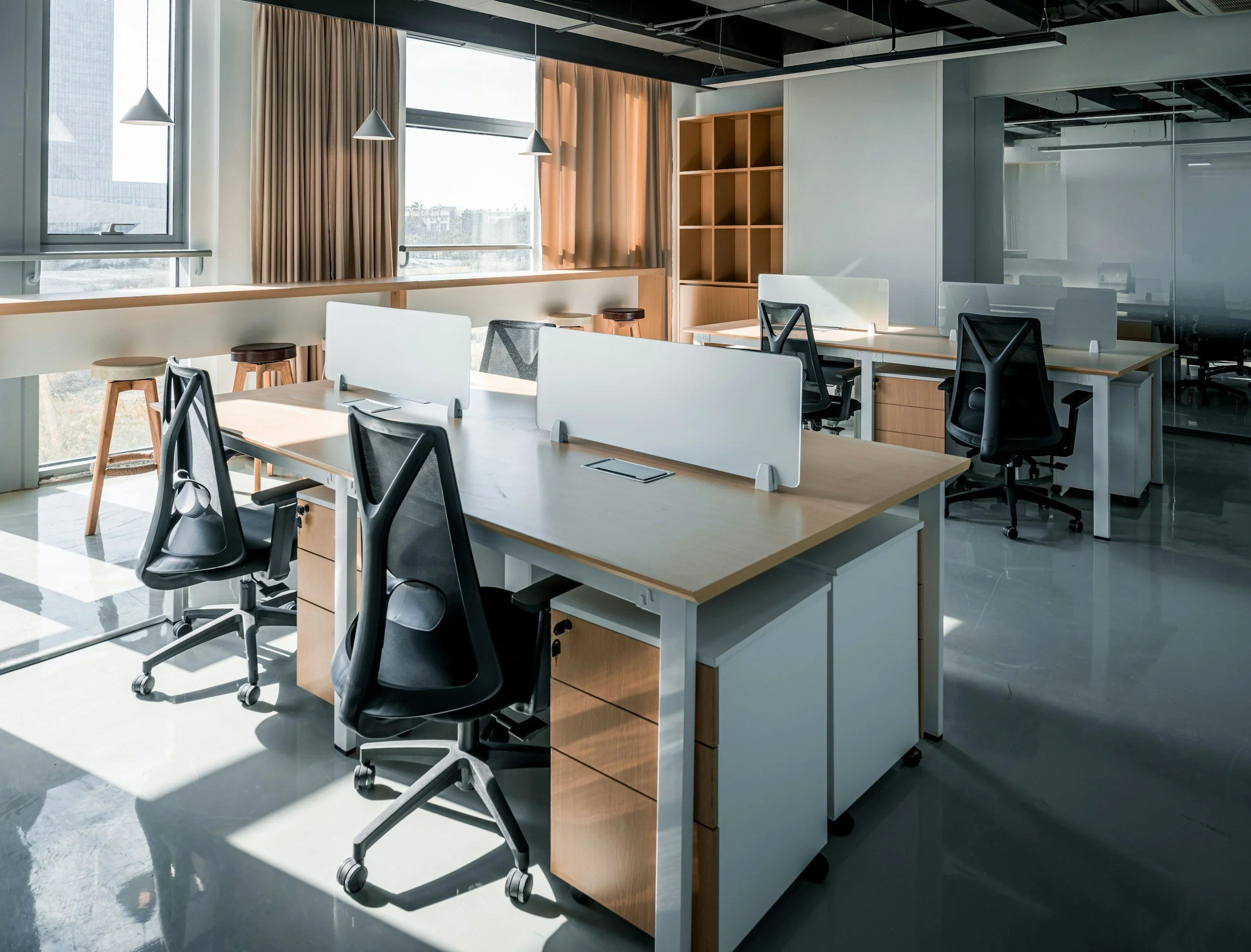 Empty modern office space with desks, chairs, and dividers, large windows with curtains, and sunlight casting shadows on the floor.