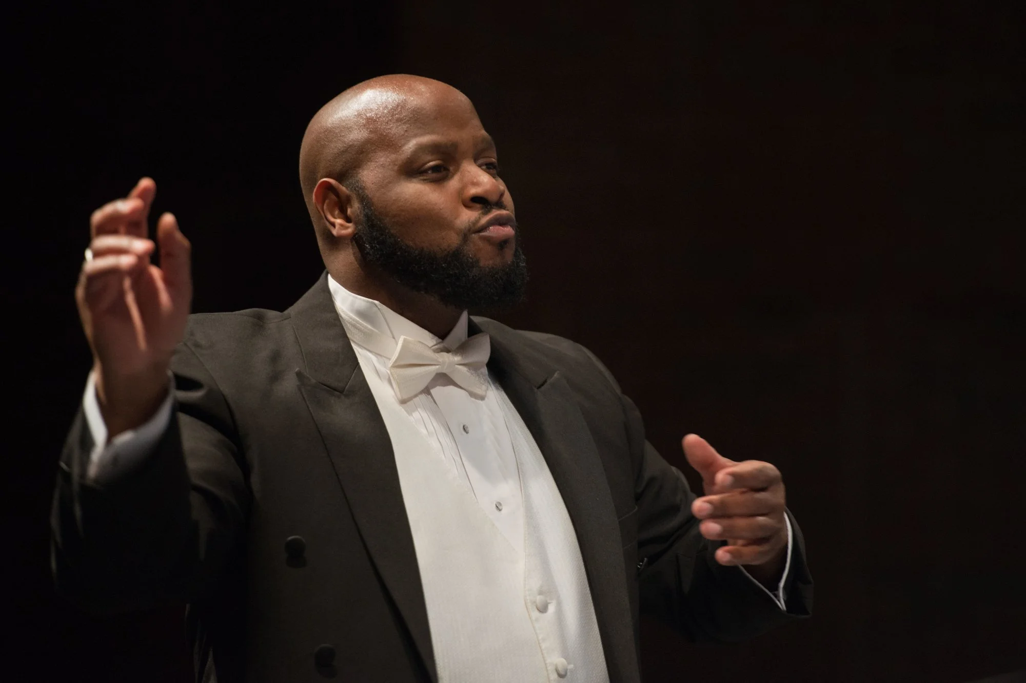 A man conducting an orchestra, dressed in a tuxedo with a bow tie, standing against a dark background.