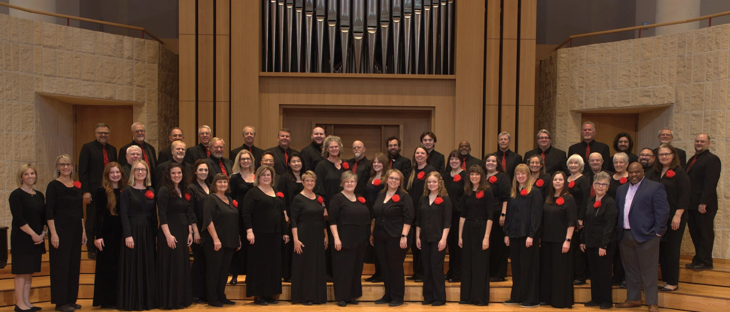 A group of men and women dressed in black with red flowers pinned to their shirts, standing on a stage in a concert hall with a wood-paneled organ in the background.
