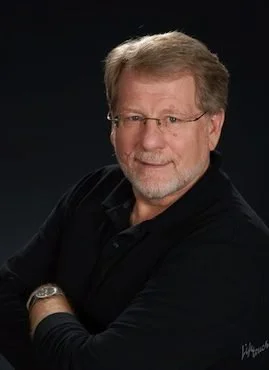 A portrait of a middle-aged man with glasses, gray hair, and a beard, wearing a dark jacket and smiling against a dark background.
