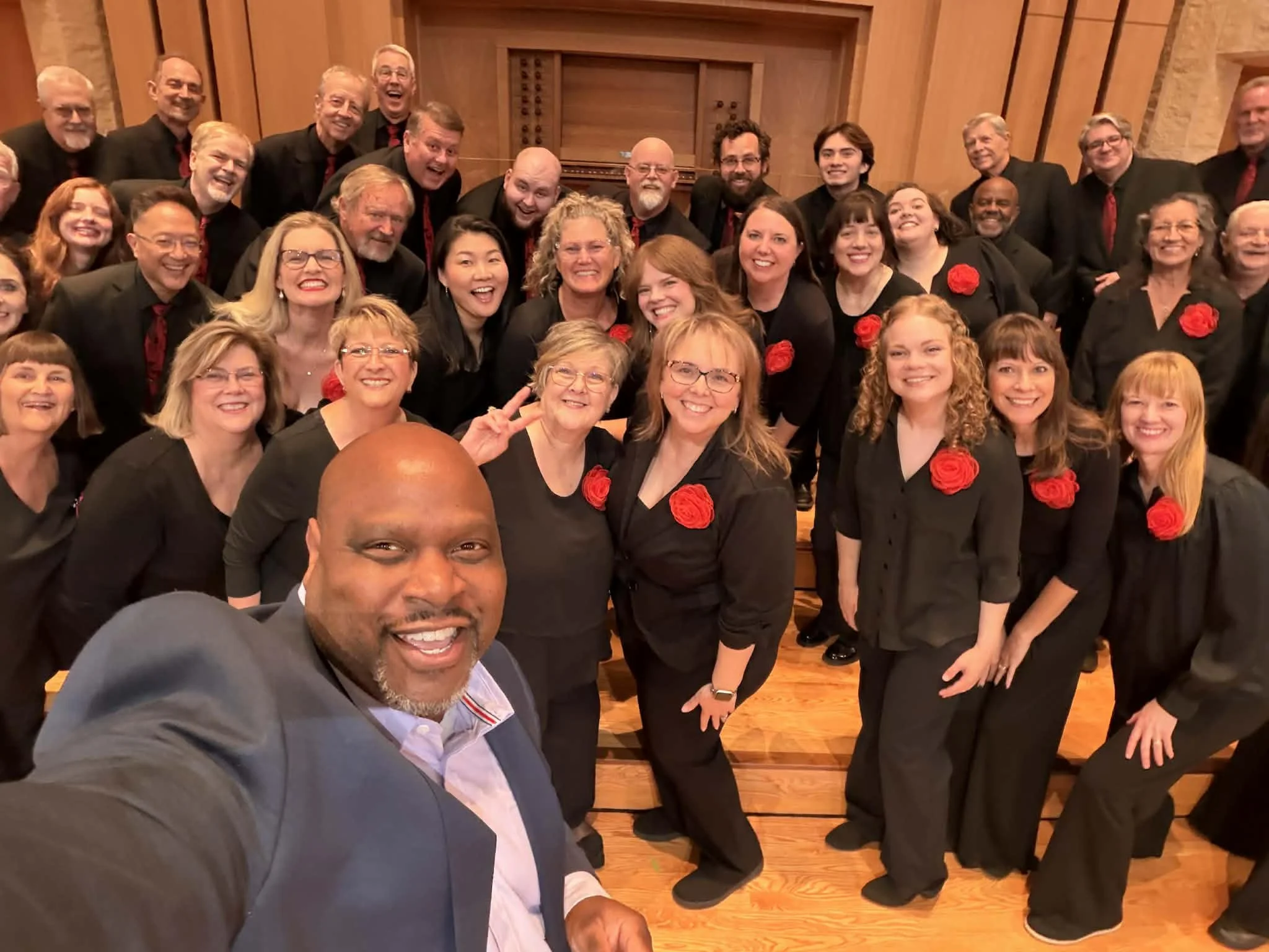 A group of smiling people dressed in formal black attire with red roses and ties, taking a selfie in a wooden room with a piano in the background.
